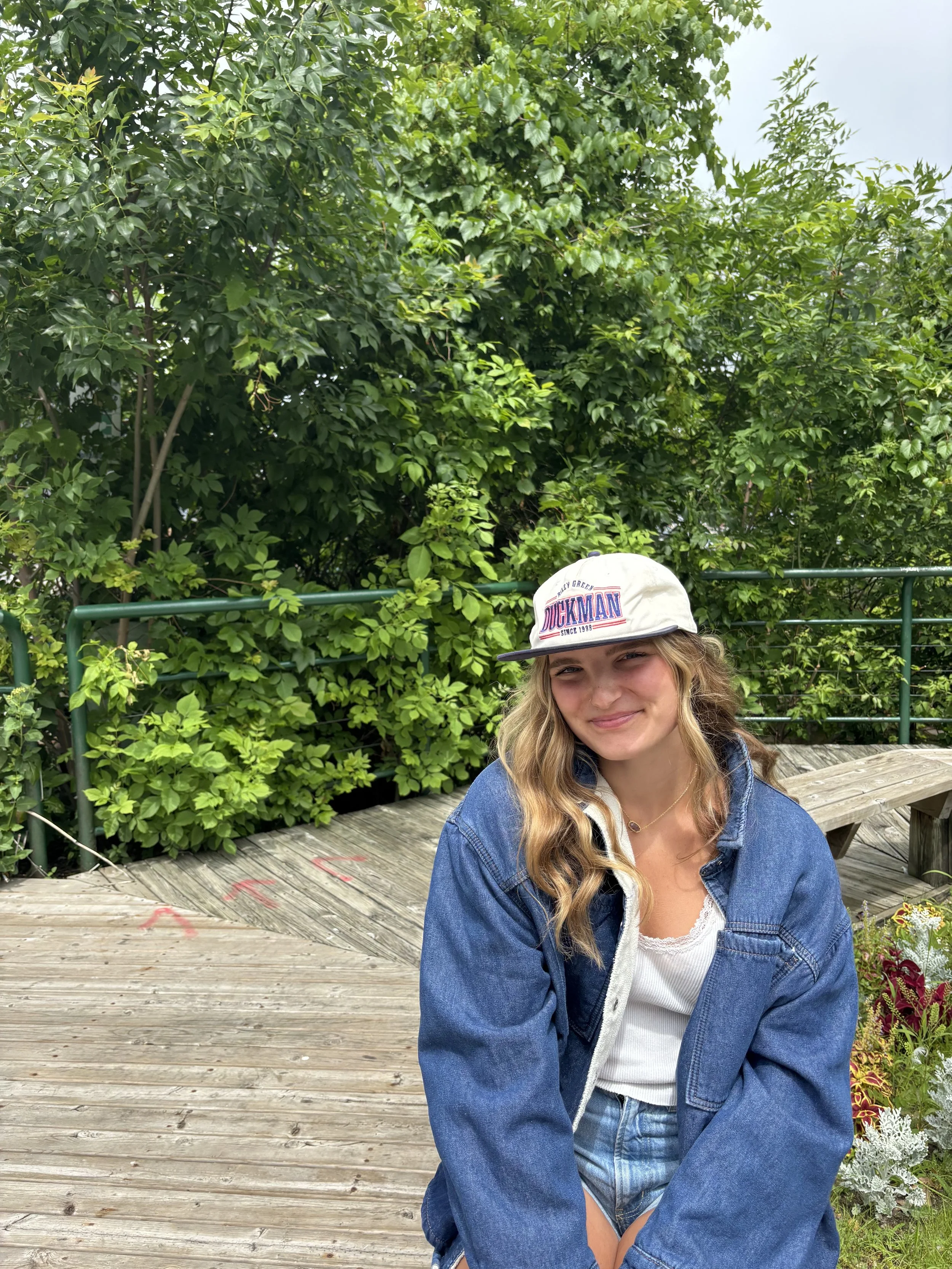 A young woman with long blonde hair wearing a white cap, a denim jacket, and a white shirt sitting on a wooden deck with lush green trees and plants in the background.