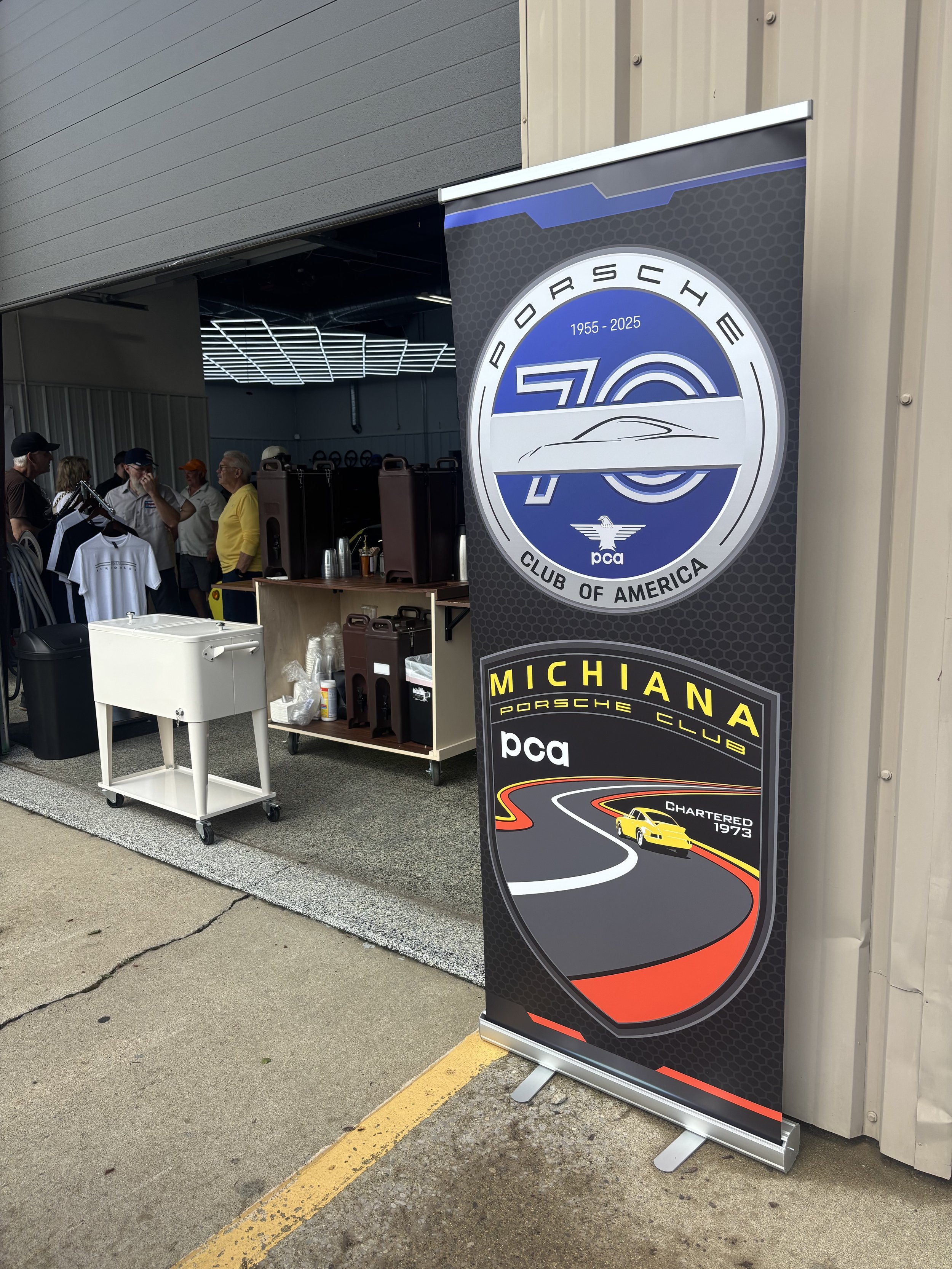 A vertical banner celebrating 70 years of Porsche Club of America, with a race car graphic and the word 'Michigan' at the bottom, in front of a metal building at an event.