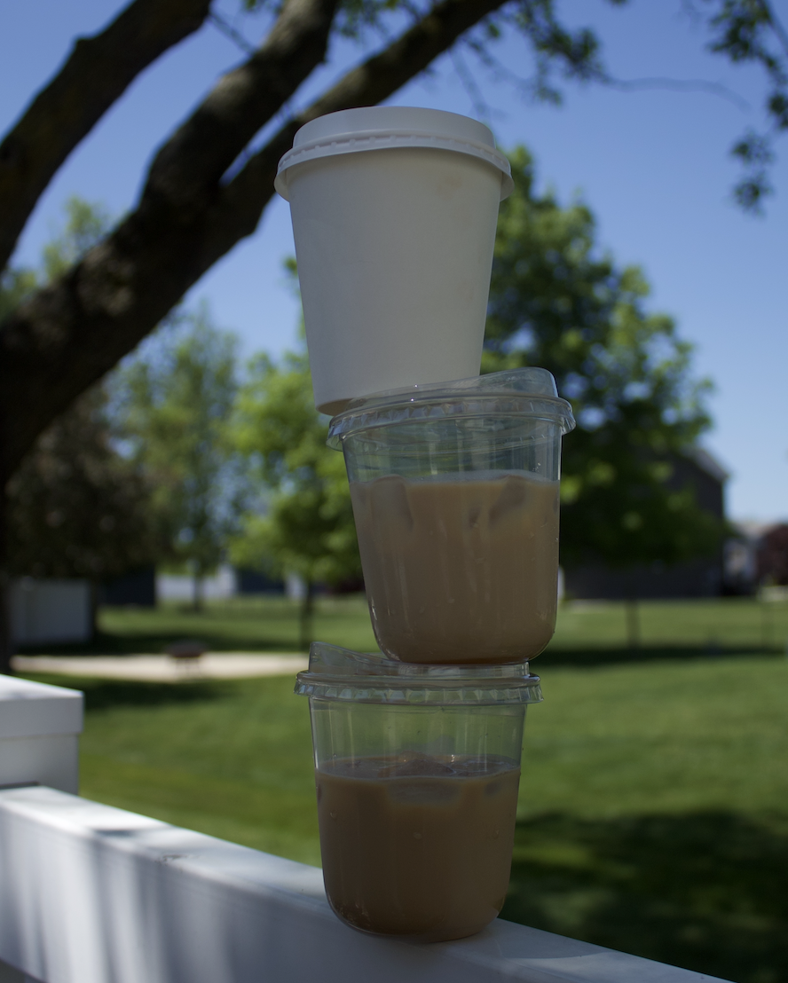 Three plastic cups of iced coffee stacked vertically with a white takeaway cup on top, outdoors on a sunny day with trees and a grassy background.