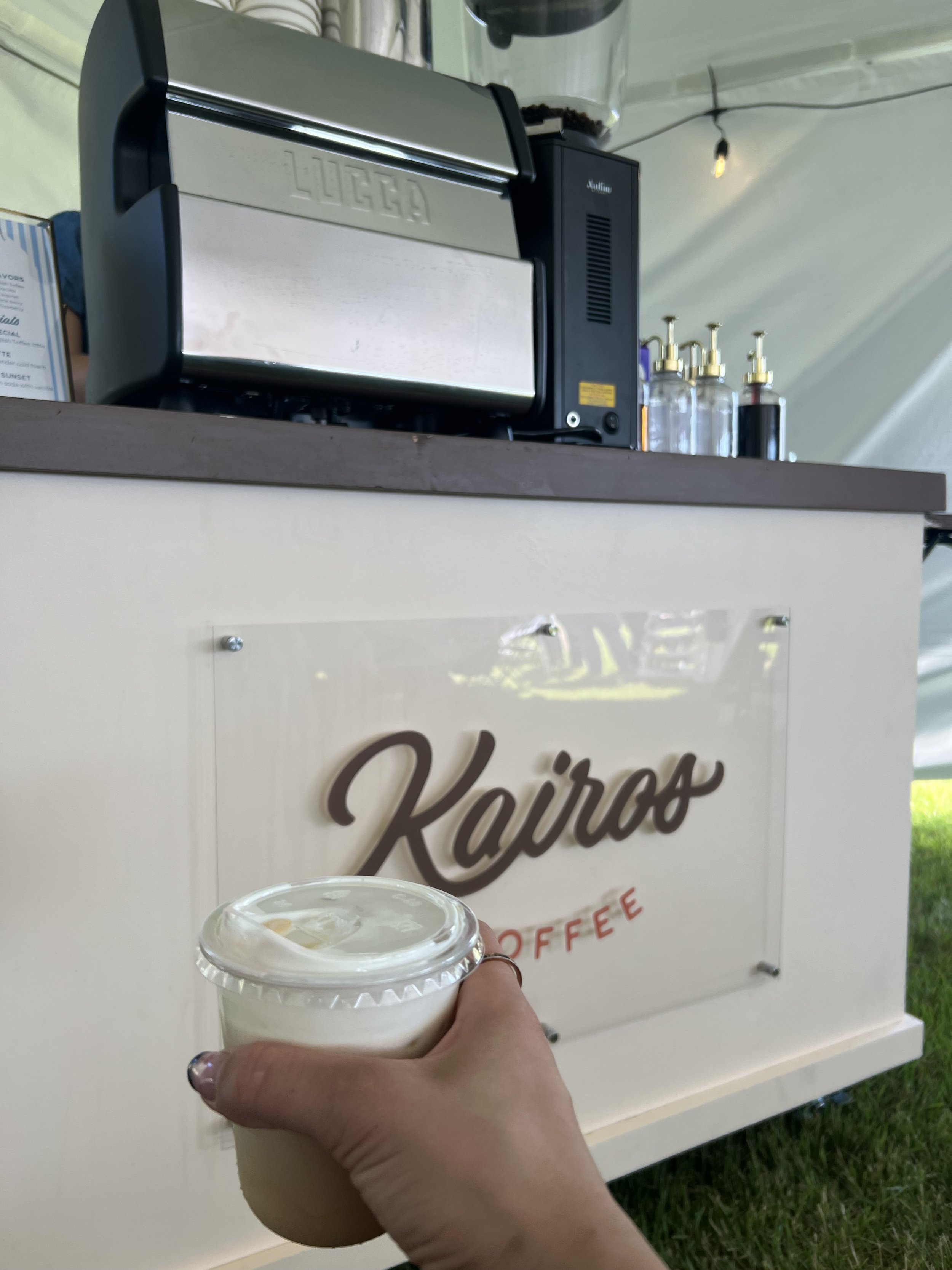 A hand holding a plastic cup with a beverage in front of a coffee kiosk with the word 'Kairo' on it. The kiosk has a coffee machine and bottles of syrup or flavoring on top.