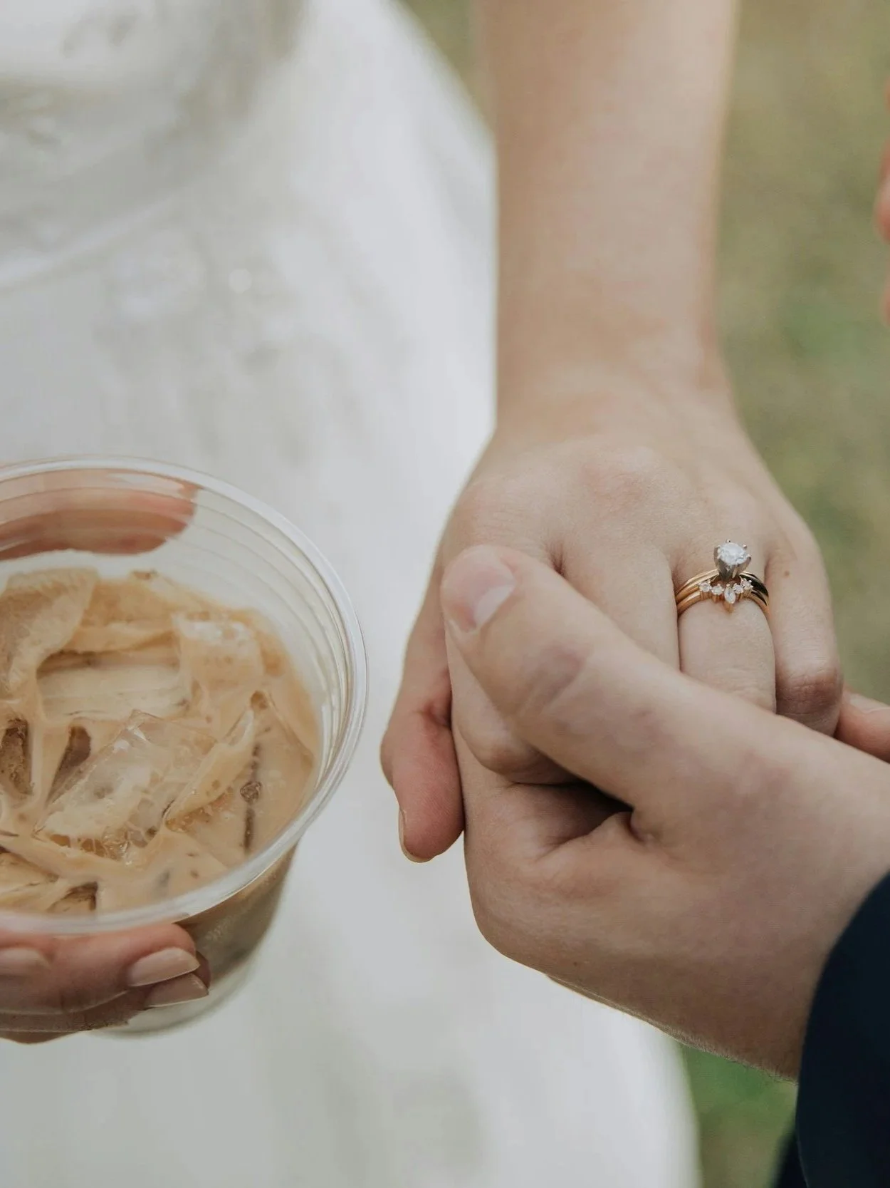 A person holding hands with another person, showing a ring with a large central gemstone. The other person is holding a cup filled with iced coffee.