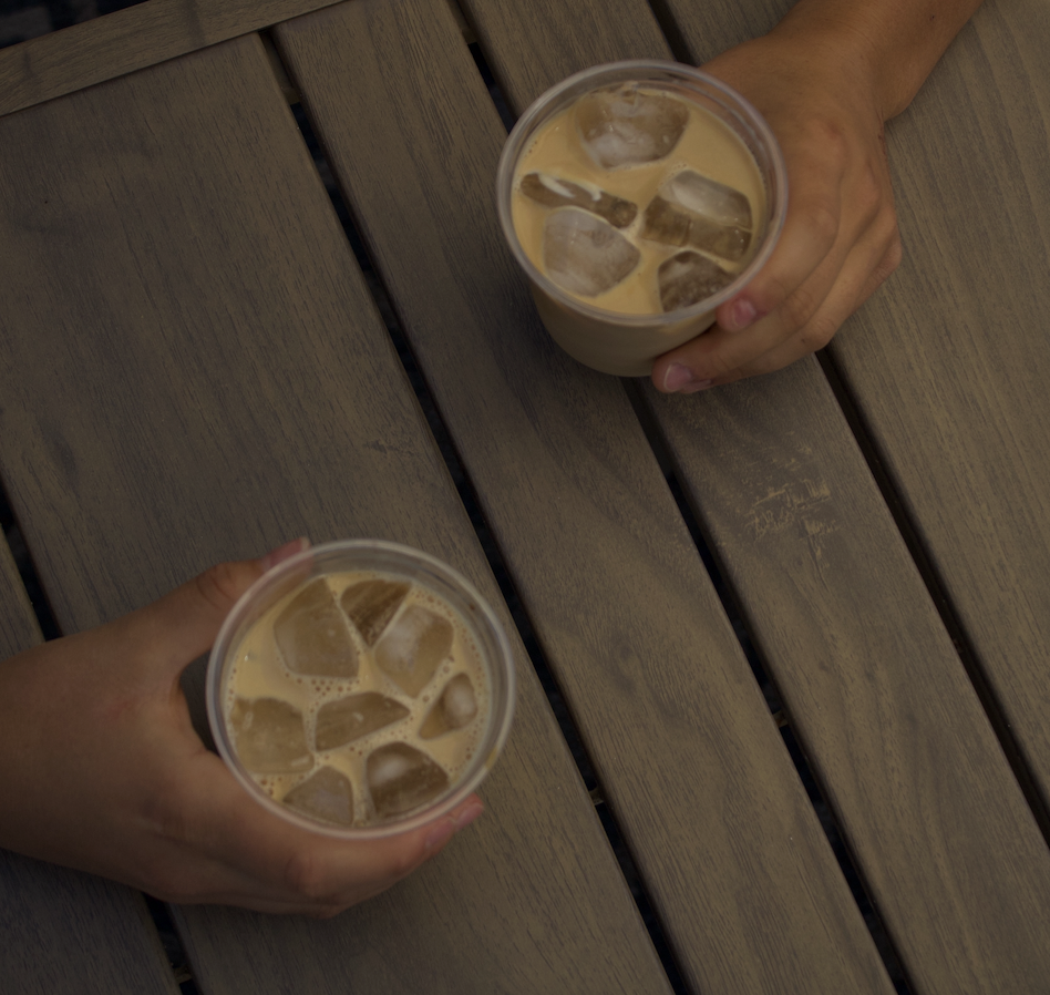 Two hands holding plastic cups with iced coffee or tea, viewed from above on a wooden table.