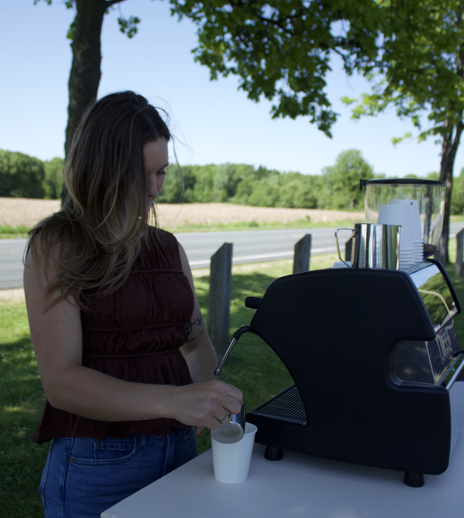 A woman is preparing a beverage in an outdoor setting with a coffee or espresso machine on a portable table. She is holding a utensil over a white cup, with trees and a road in the background on a sunny day.