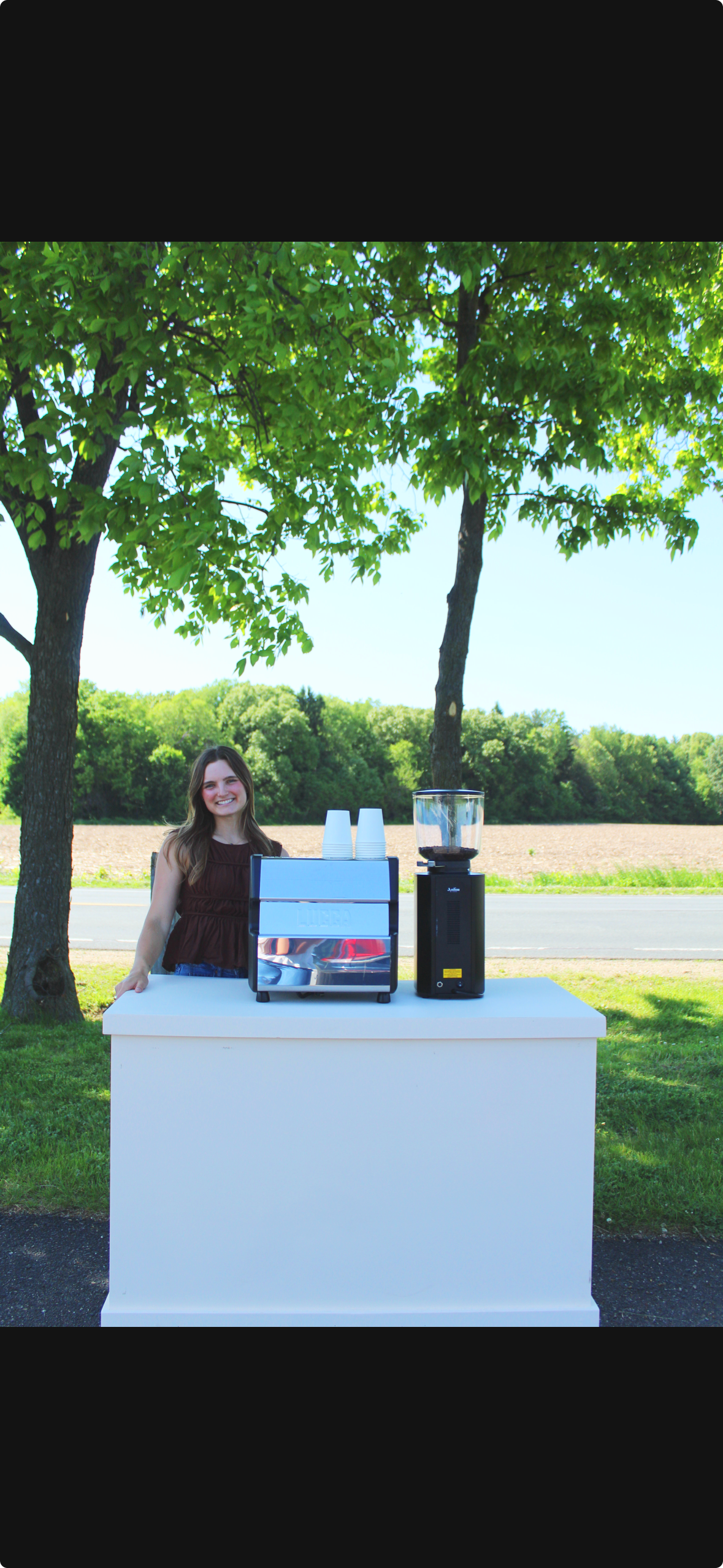 A young woman with long brown hair smiling standing behind a white counter with an espresso machine and a coffee grinder outdoors under a large leafy tree, with a field and trees in the background.