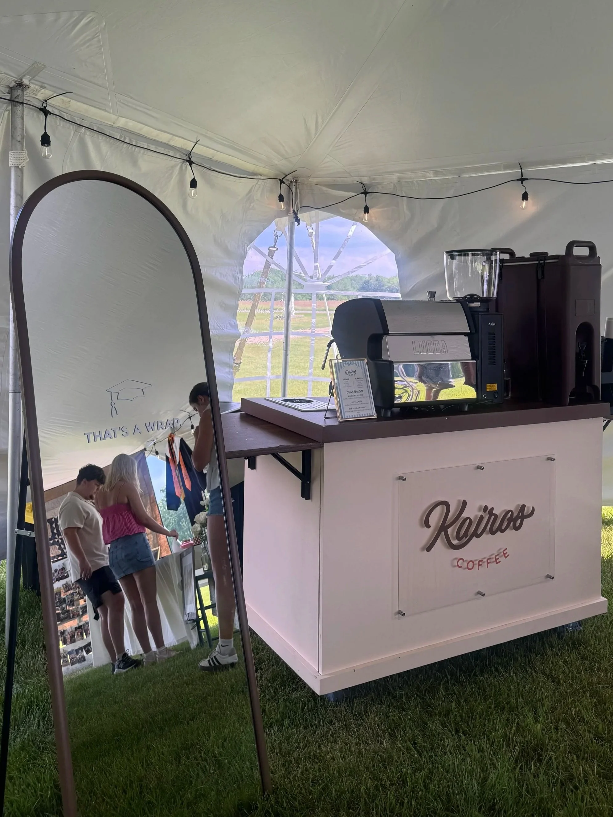 A tent setup with a coffee stand that reads 'Kairo Coffee' on the front. Inside the tent, there is a coffee machine and a small sign with menu information. Several people are gathered near the stand, and a large mirror on a stand is visible on the left side of the image. String lights hang from the tent's ceiling, and a windmill is visible through the tent opening in the background.