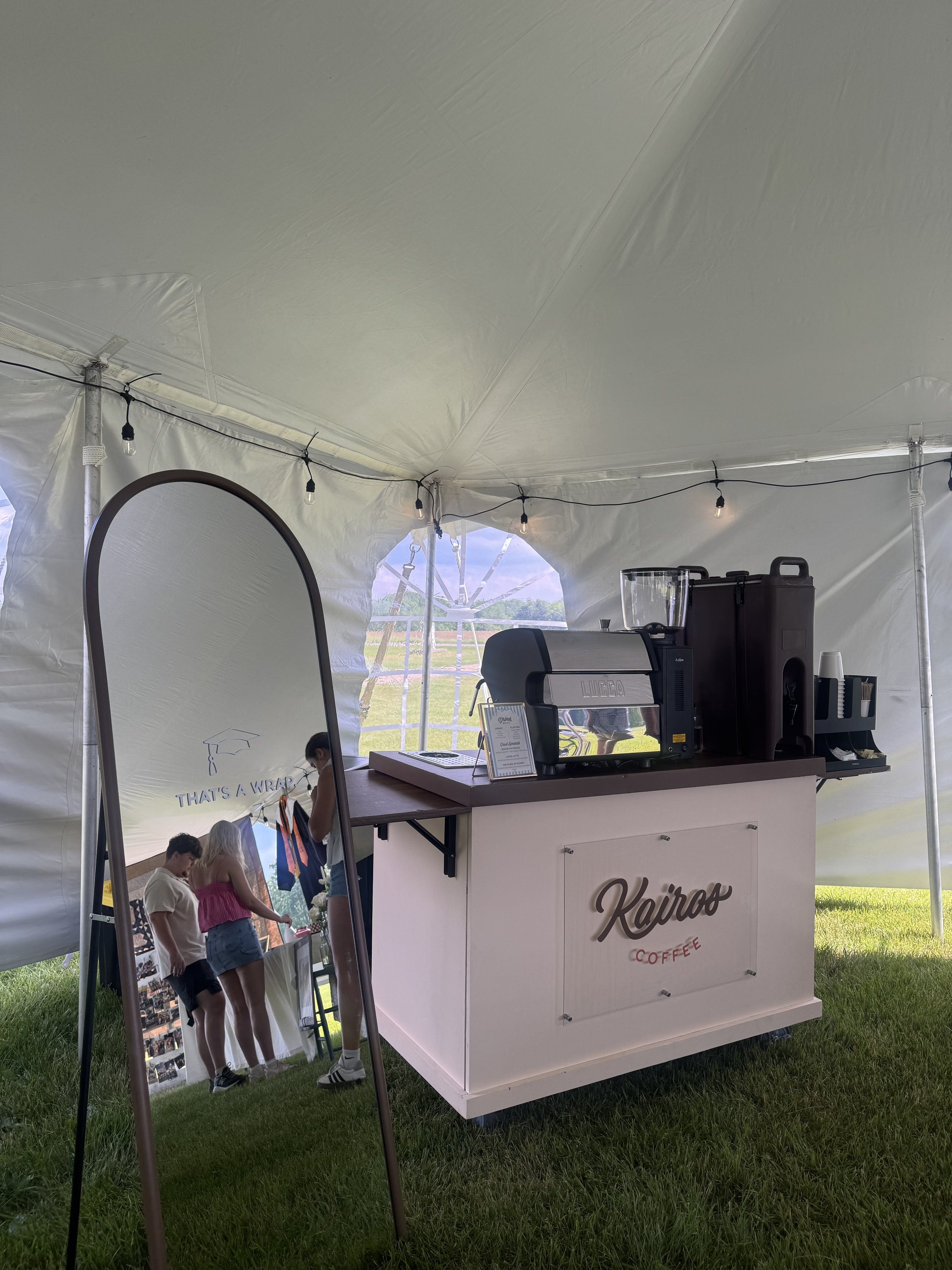 A white coffee stand with the name 'Kairo' on the front, inside a large white event tent. The stand has a coffee grinder, cups, and a small menu. A mirror and a sidewalk chalkboard with the message 'THAT'S A WRAP' are visible. Outside the tent, a few
