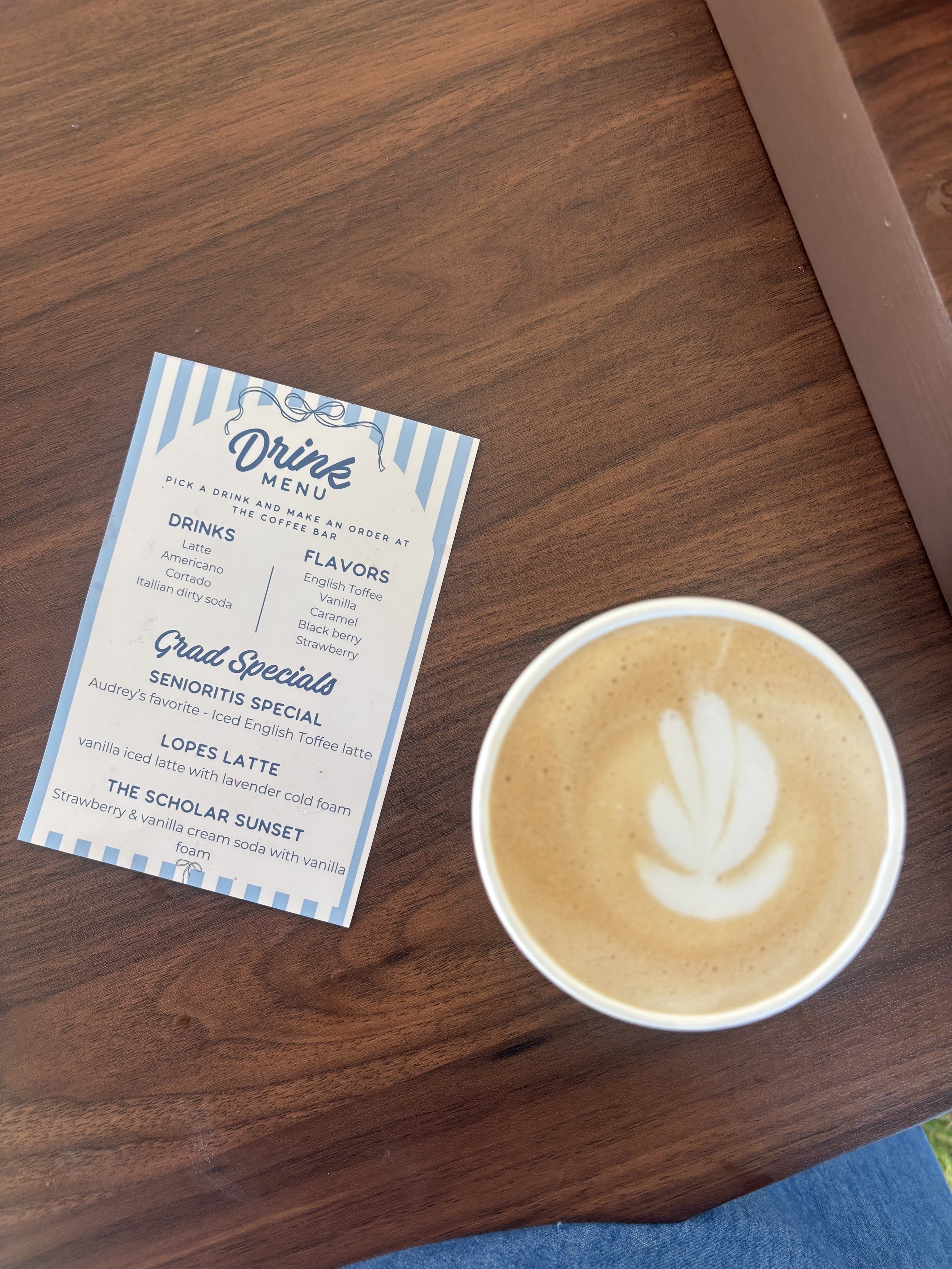 A wooden table with a drink in a paper cup showing a swirl of cream on top, and a menu with a blue and white striped border and list of drink options.