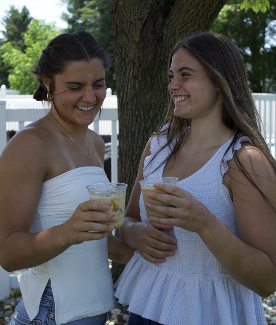Two young women smiling and holding drinks outdoors near a tree with a white fence in the background.