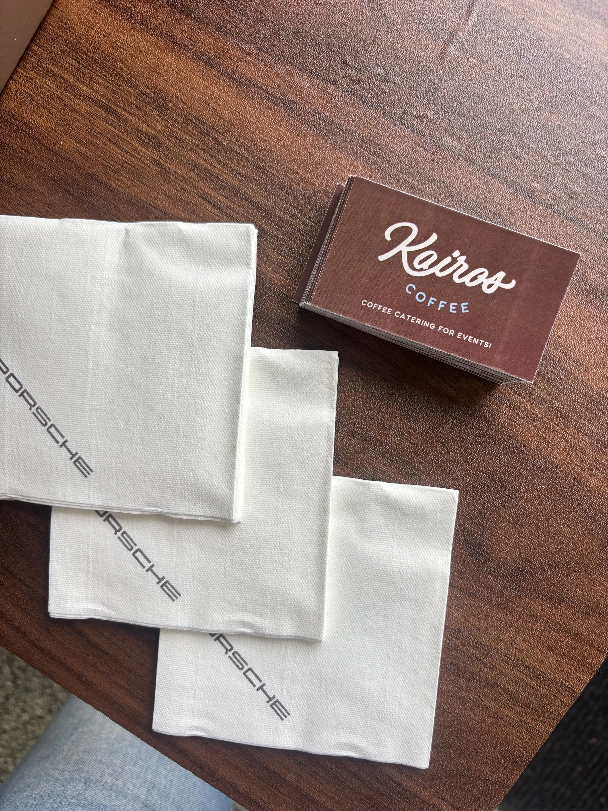 A business card for Keiros Coffee on a wooden table, with white Porsche-branded napkins nearby.