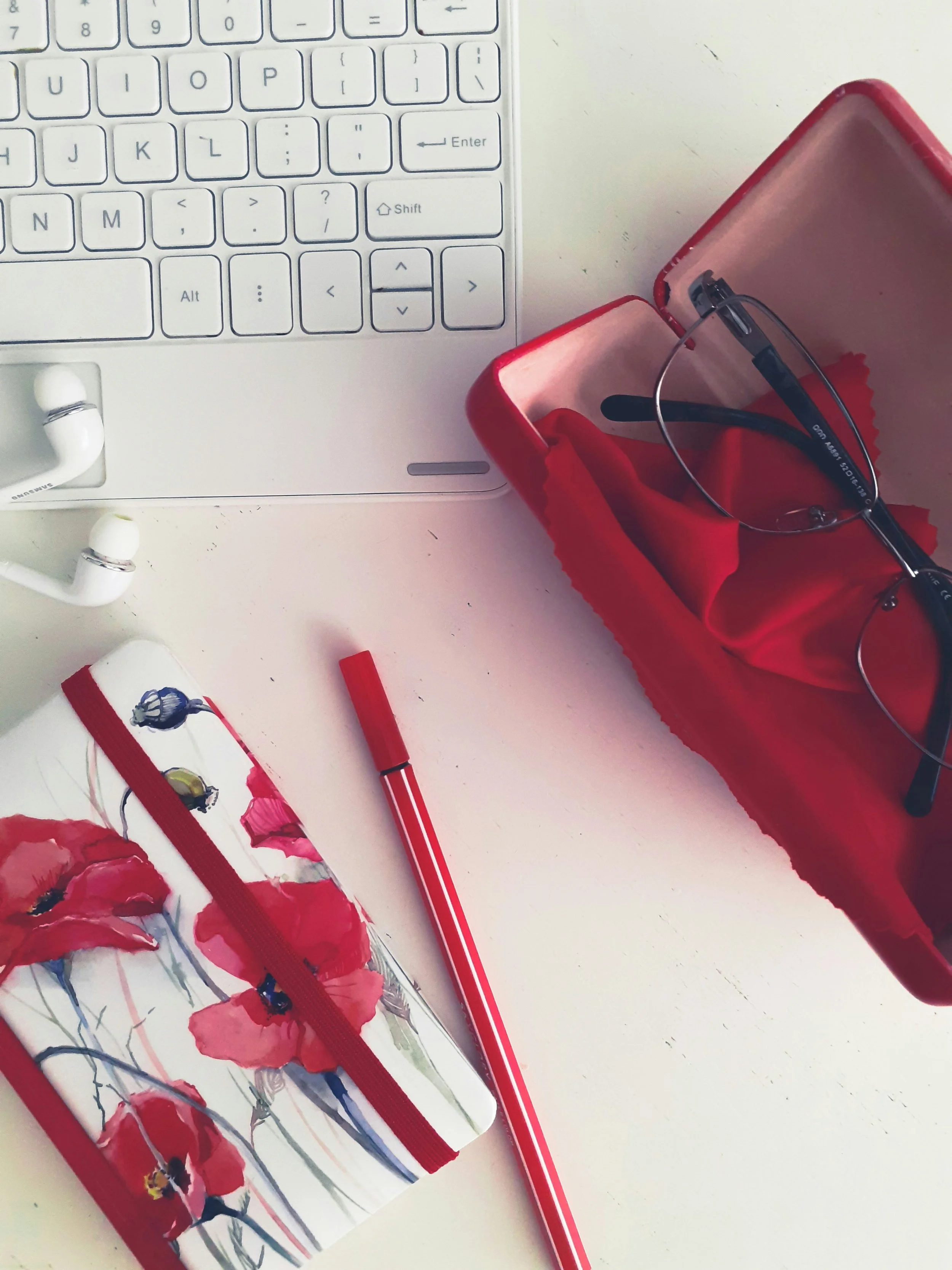 A white computer keyboard, a pair of white earbuds, a red and white striped pen, and a red pencil case containing glasses lie on a white desk.