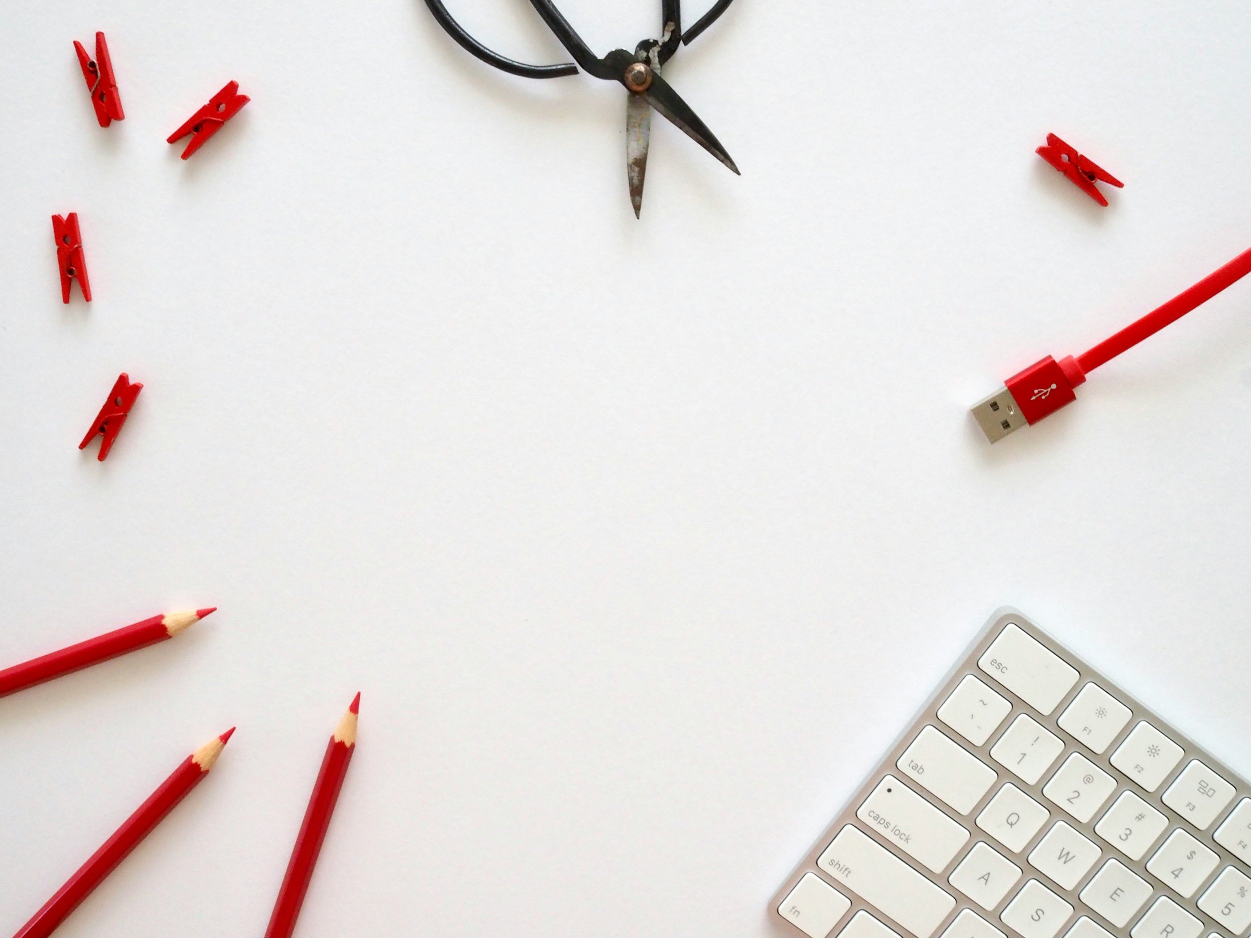 Close-up of a white desk with scattered red pencils, red clothespins, a partially visible white keyboard, a white computer mouse, a pair of vintage black scissors, a red USB cable, and a portion of a white keyboard.