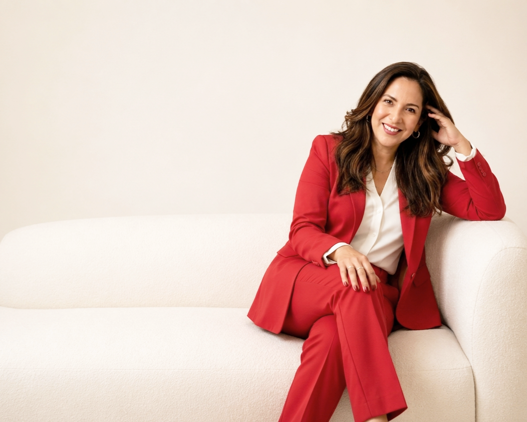 A woman in a red suit sitting on a white couch, smiling, against a plain beige background.