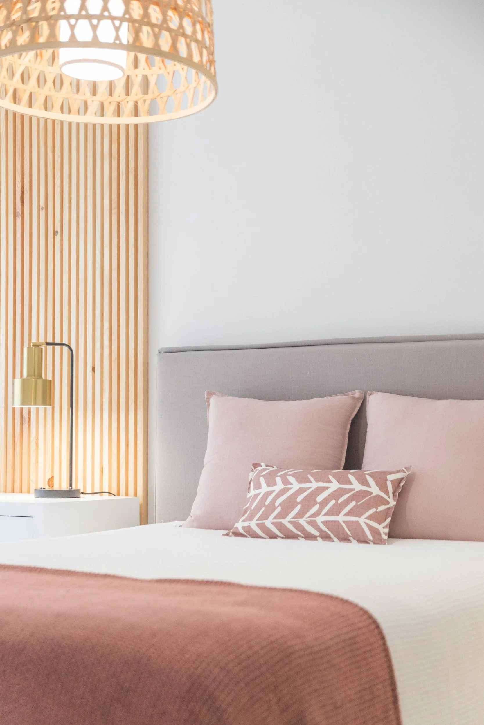 A bedroom with a bed, pink pillows, a pink blanket, a bedside table with a gold lamp, and a wooden lattice headboard with a modern hanging light fixture.