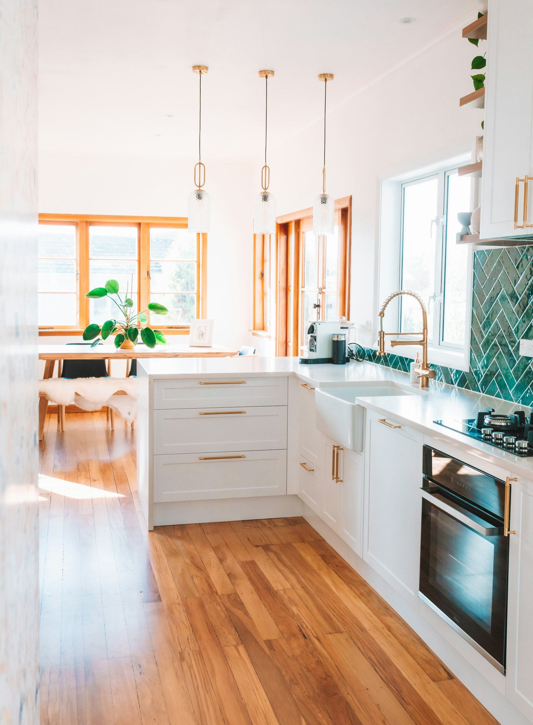 Bright kitchen with white cabinets, gold handles, a gold faucet, wooden floors, large windows, hanging pendant lights, and a countertop with kitchen appliances and decor.