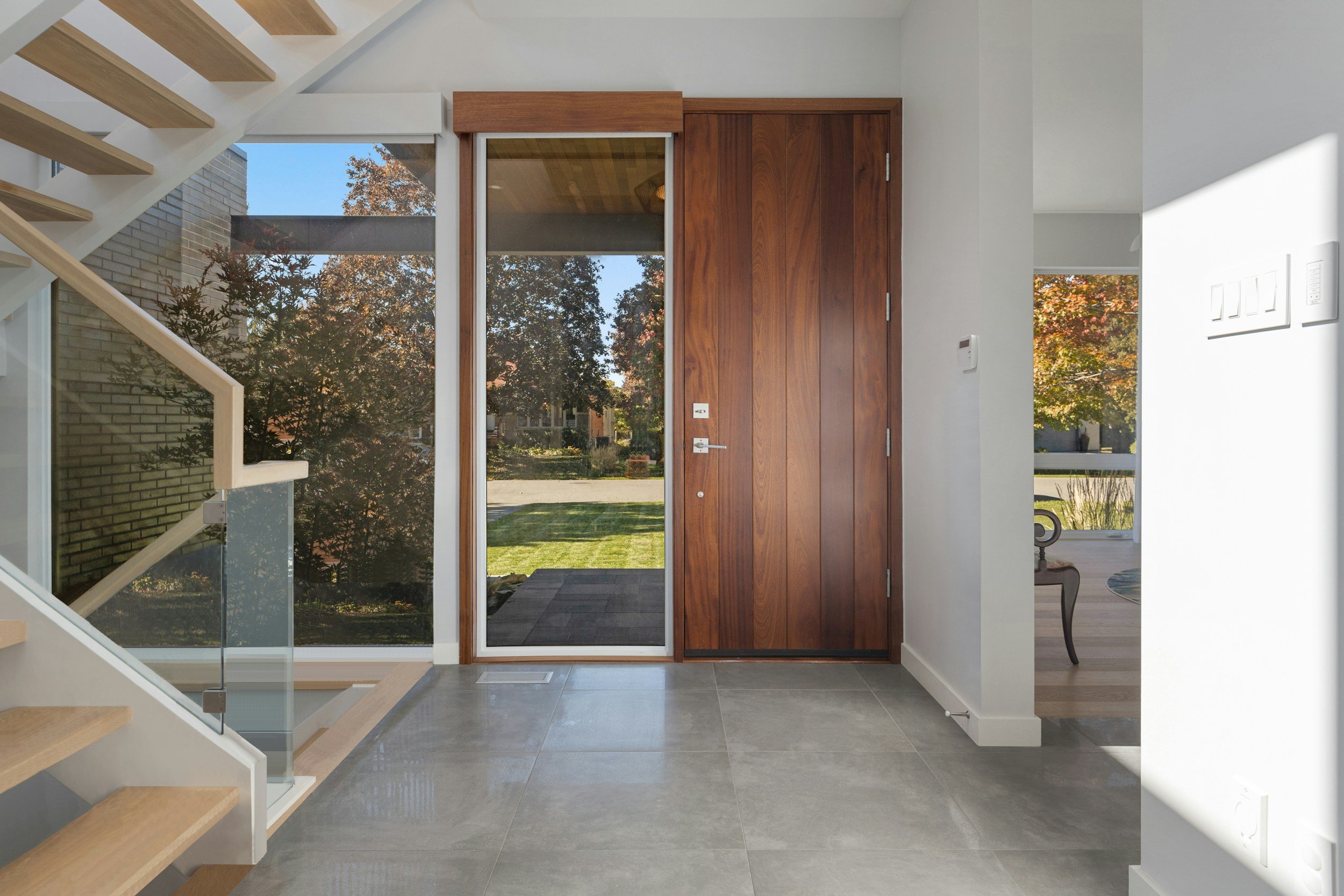 Modern entrance with a wooden door, glass panel, staircase, and large windows showing trees outside.