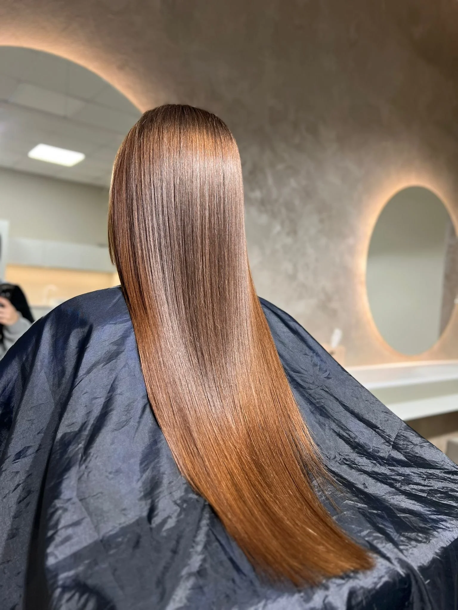 Person with straight, shiny, reddish-brown hair sitting in a salon chair, facing away from the mirror, with a large oval mirror on the wall behind them.