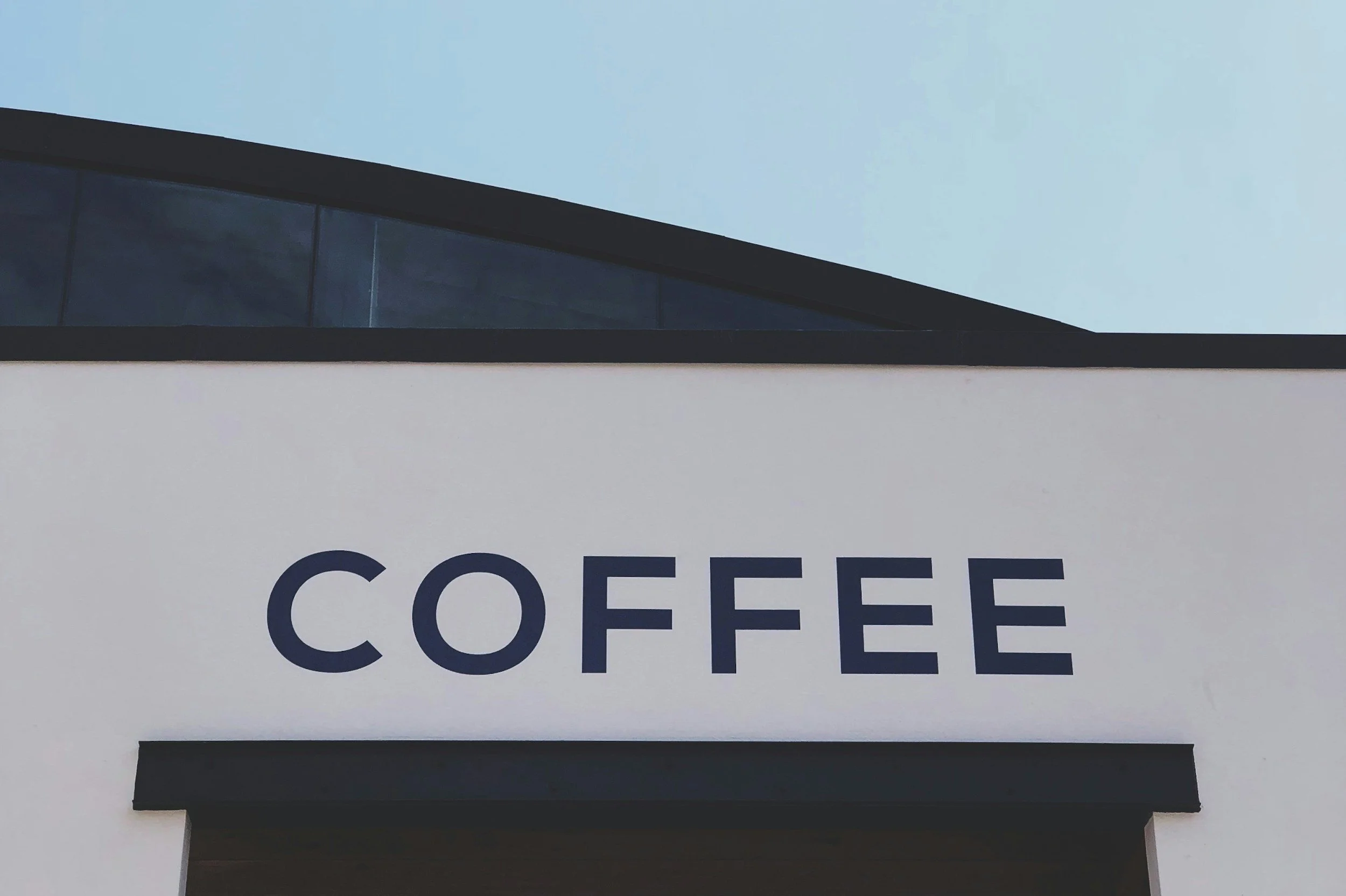 Close-up of a commercial building with the word 'COFFEE' written on the sign, partial view of the facade and glass window at the top, against a clear sky.