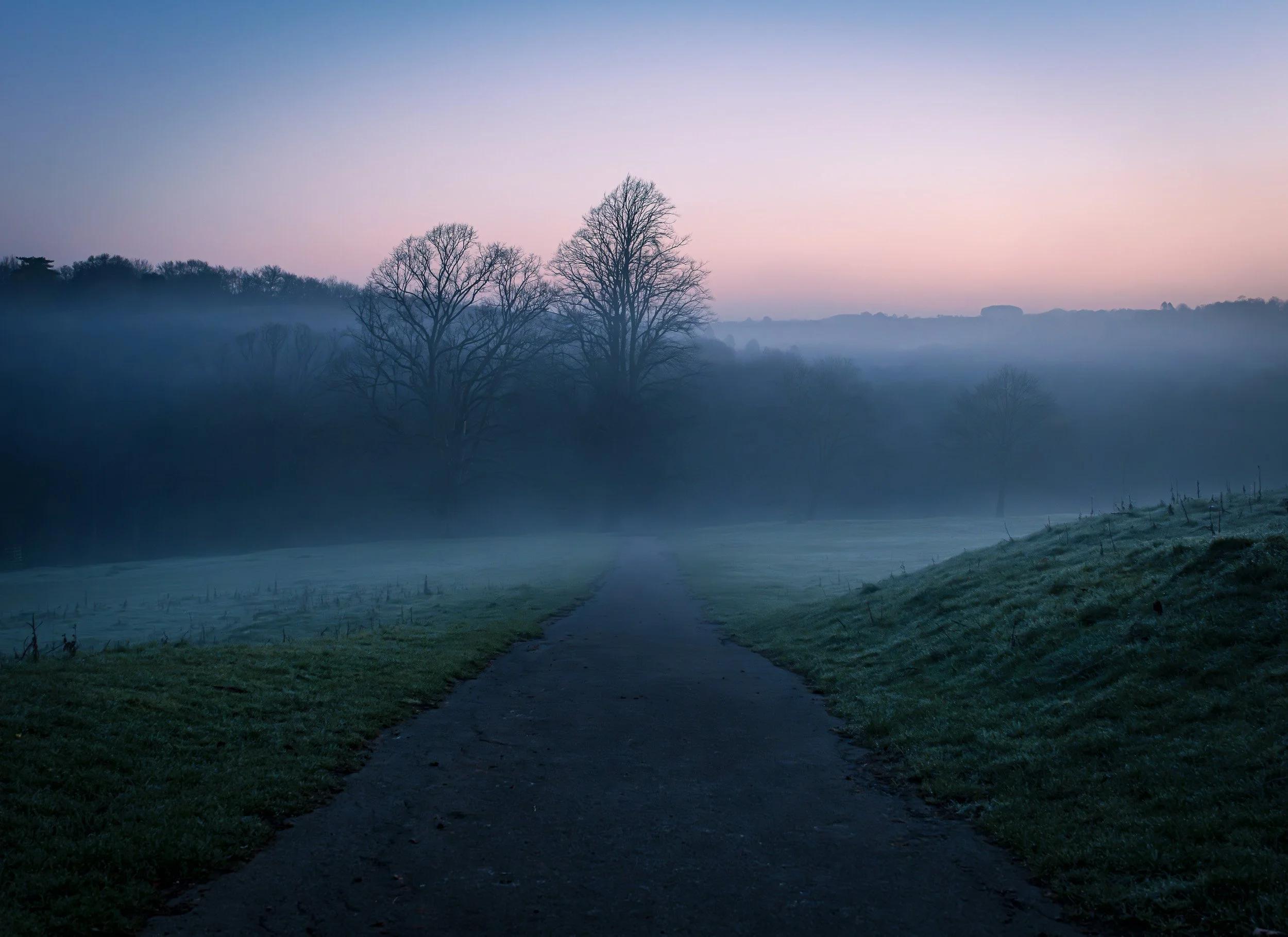 A scenic landscape during dusk or dawn with a winding gravel path leading towards a body of water, surrounded by grassy fields. Bare trees are visible, and mist or fog hovers low over the ground with a pastel-colored sky in the background.
