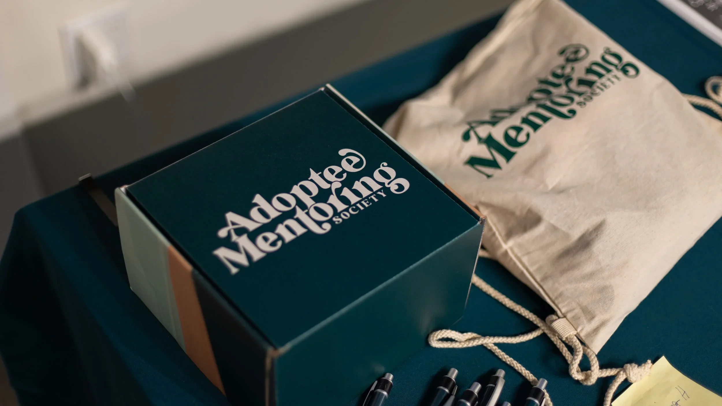 A box and a paper bag both labeled 'Adoptee Mentoring Society' placed on a dark blue table with pens and a yellow sticky note nearby.