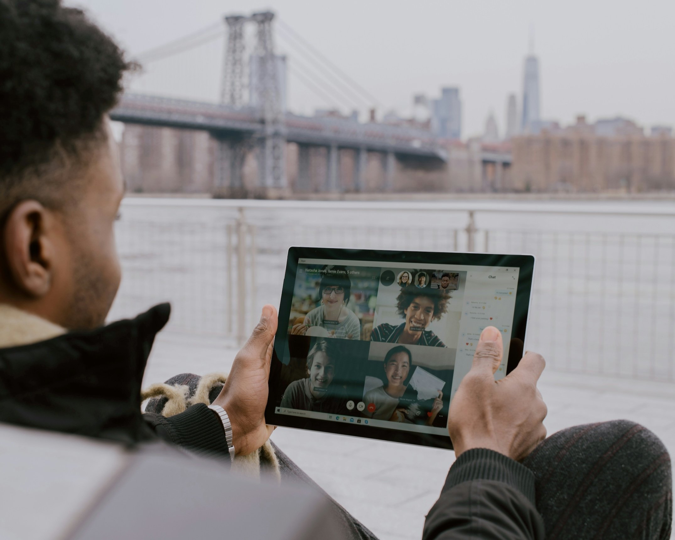 Person holding a tablet displaying a video call with multiple smiling participants, outdoors near a body of water with a bridge and skyline in the background.