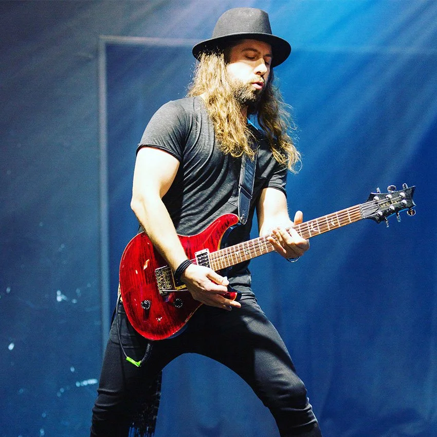 Male guitarist with long hair and beard, wearing a black hat and black t-shirt, playing a red electric guitar on stage.