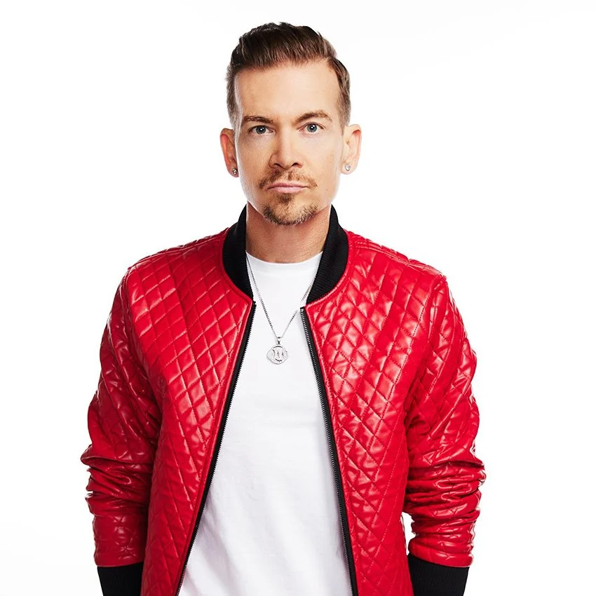 A man with short brown hair,goatee, and earrings wearing a red quilted leather jacket over a white T-shirt, with a silver necklace, standing against a white background.