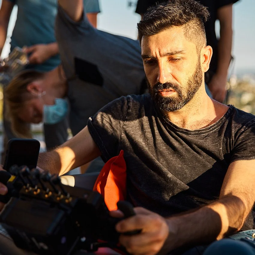 A man with a beard in a black shirt looks at his camera, with a woman wearing a face mask and medical scrubs in the background.