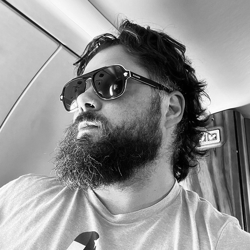 A man with a beard and mustache wearing sunglasses, a nose ring, and a T-shirt, sitting inside an airplane.
