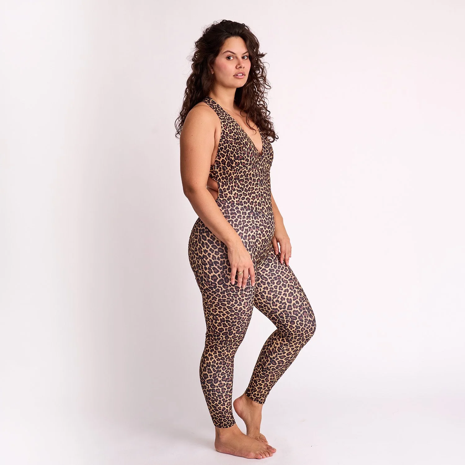 A woman with curly brown hair is standing barefoot against a plain white background, wearing a matching sleeveless leopard print top and leggings.
