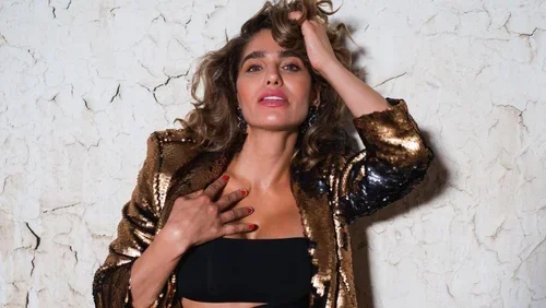 A woman with curly hair, wearing a black top and a shiny, gold jacket, standing against a cracked white wall.