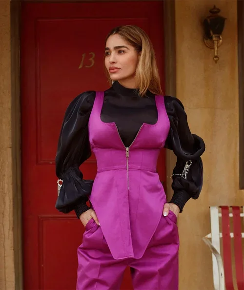 A woman with shoulder-length blonde hair, wearing a black top with puffy sleeves layered under a vibrant pink vest with a front zipper, standing outside in front of a red door numbered 13 and a beige wall with a vintage wall lamp.