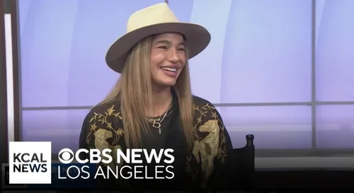 A woman with long blonde hair wearing a beige hat and a black sweater with gold patterns, smiling during an interview on CBS News in Los Angeles.