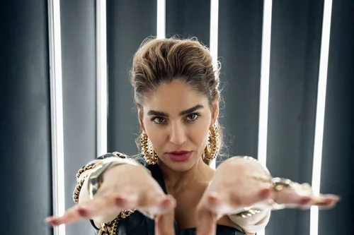 Woman with dark hair, wearing gold earrings, holding out her hands toward the camera, with vertical light bars in the background.