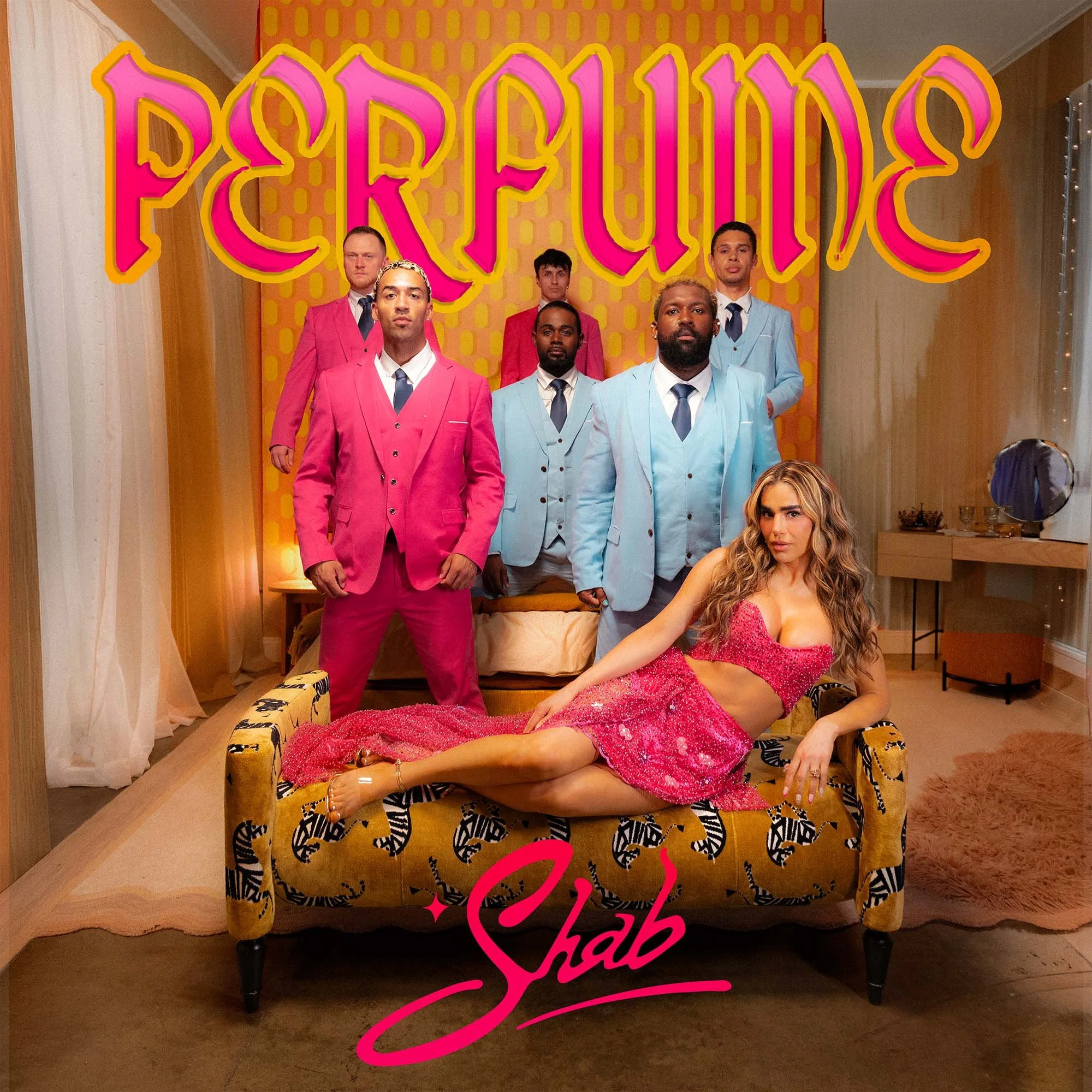 Group of five men in colorful suits standing behind a woman in a pink dress sitting on a patterned yellow sofa in a room with yellow patterned wall and large pink and yellow sign that reads 'Proudly'.