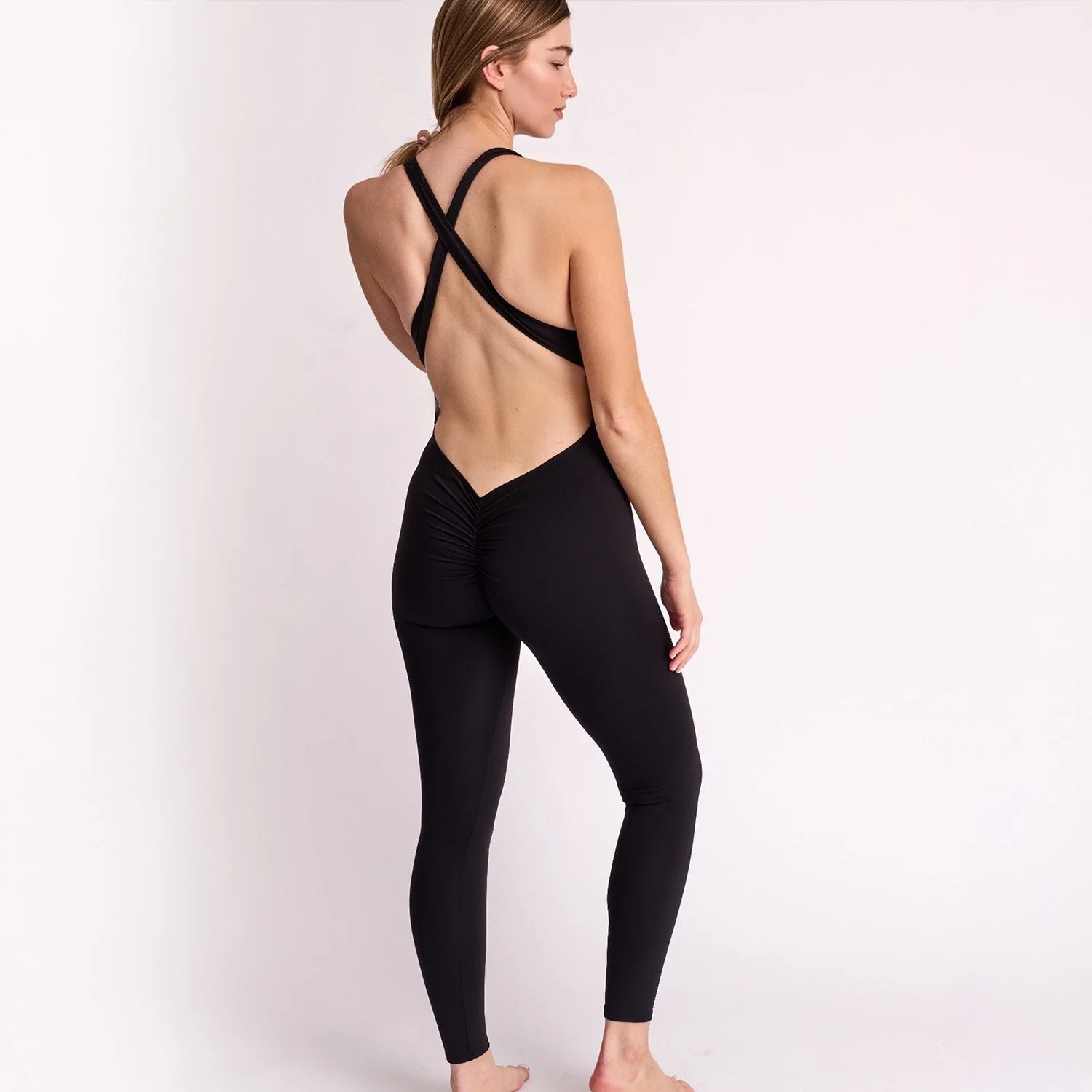 A woman with light skin and long, light brown hair tied in a ponytail, standing sideways with one hand hanging down, wearing a black backless top with crisscross straps and black fitted pants, against a plain white background.