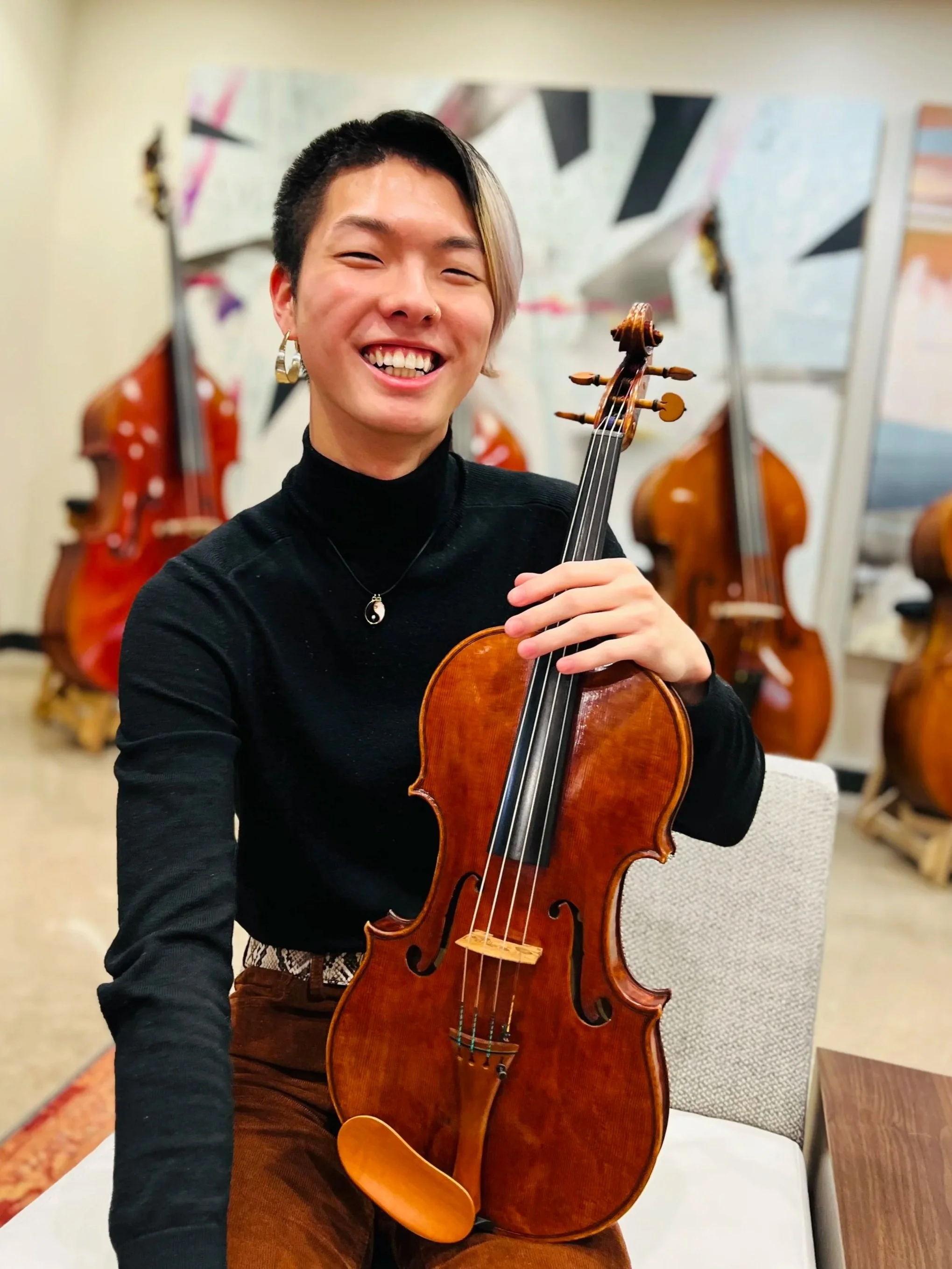 Person with short black and blonde hair smiling, holding a violin in a room with other violins in the background.