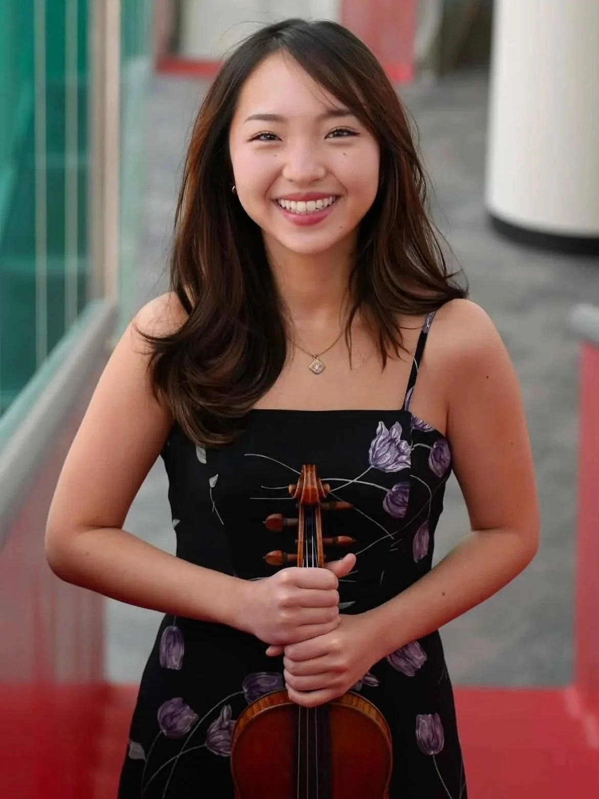 A woman with long dark hair and a bright smile holding a violin, wearing a black dress with purple floral patterns, standing outdoors with blurred background.