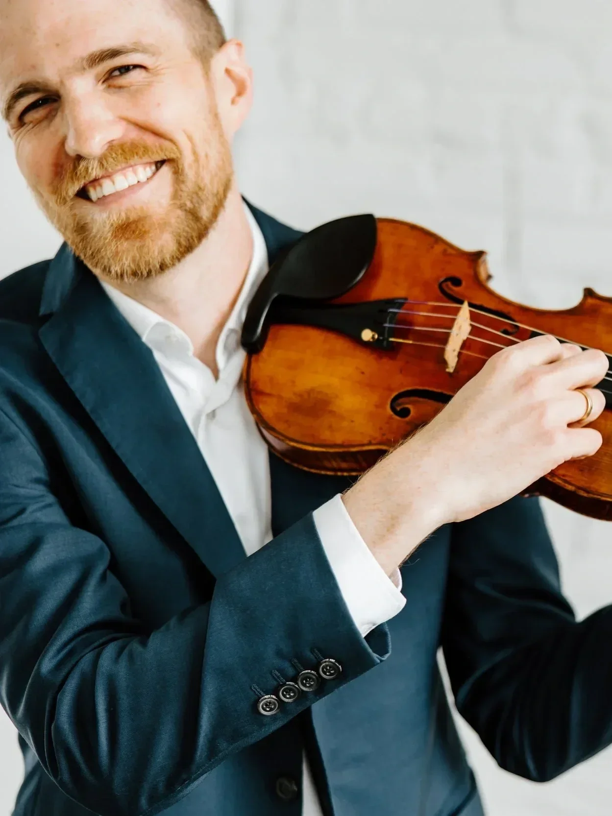 A man smiling and holding a violin, dressed in a dark suit and white shirt.