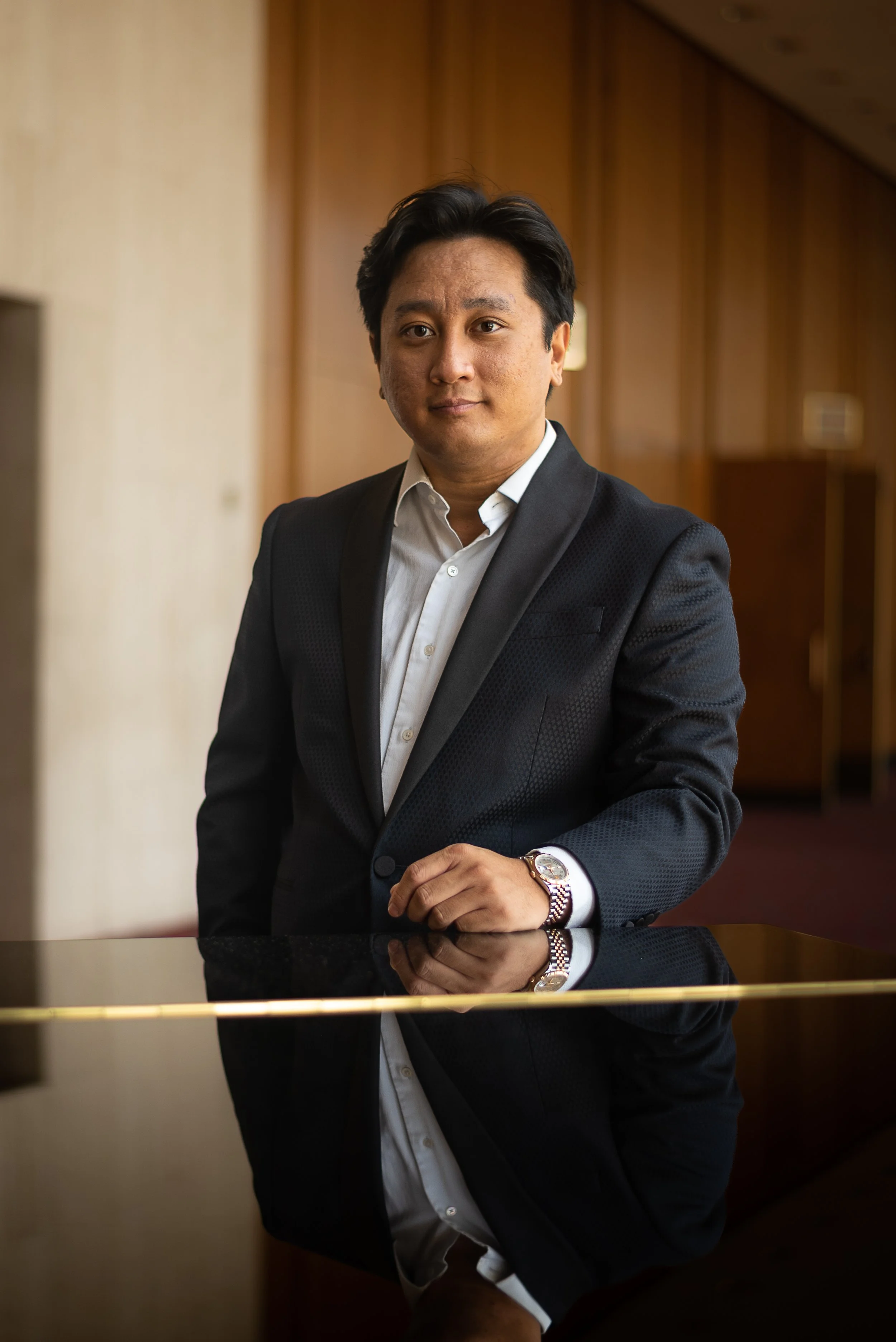 A man in a dark suit with a white shirt, standing indoors with wooden wall panels in the background. His left hand rests on a reflective black and gold surface, and he is wearing a wristwatch. The image captures a professional and confident demeanor.