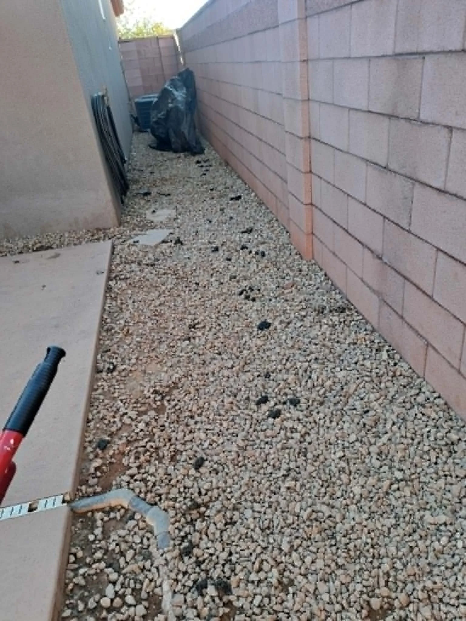 A narrow outdoor space with a gravel ground, a brick wall on the right, and a green wall on the left. There is a trash bag and a plastic storage bin in the back corner, with some bricks and a gardening tool nearby.