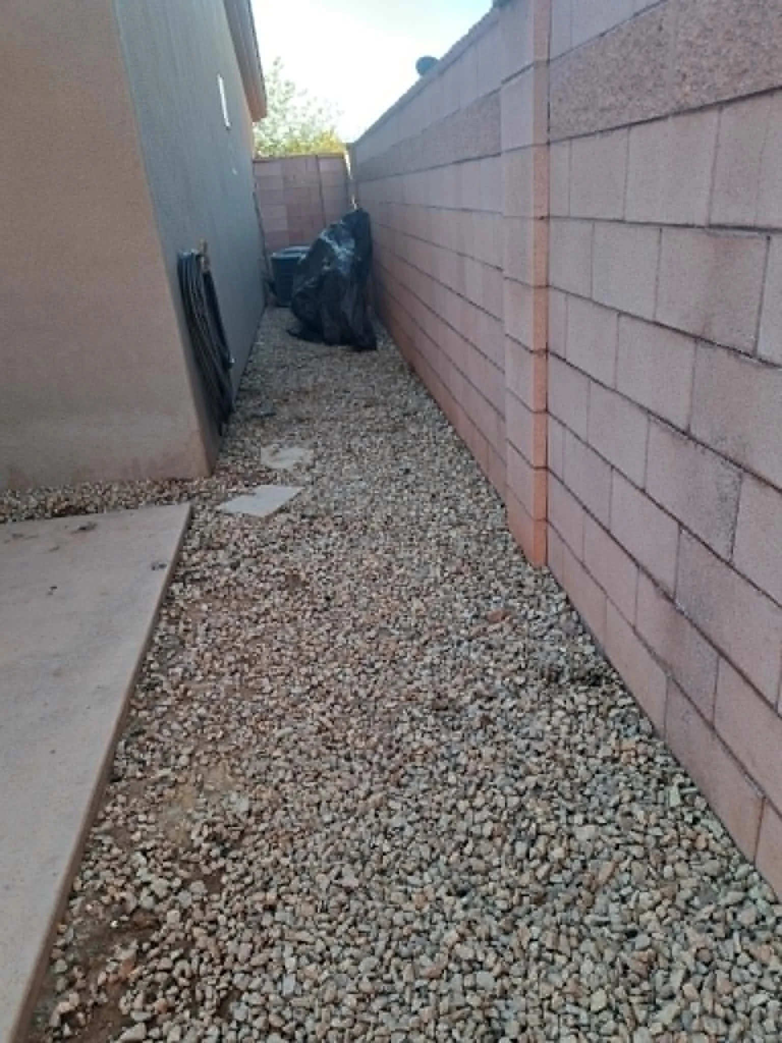 Side yard with gravel ground, concrete slab, brick wall on the right, gray wall on the left, and trash bags in the corner.