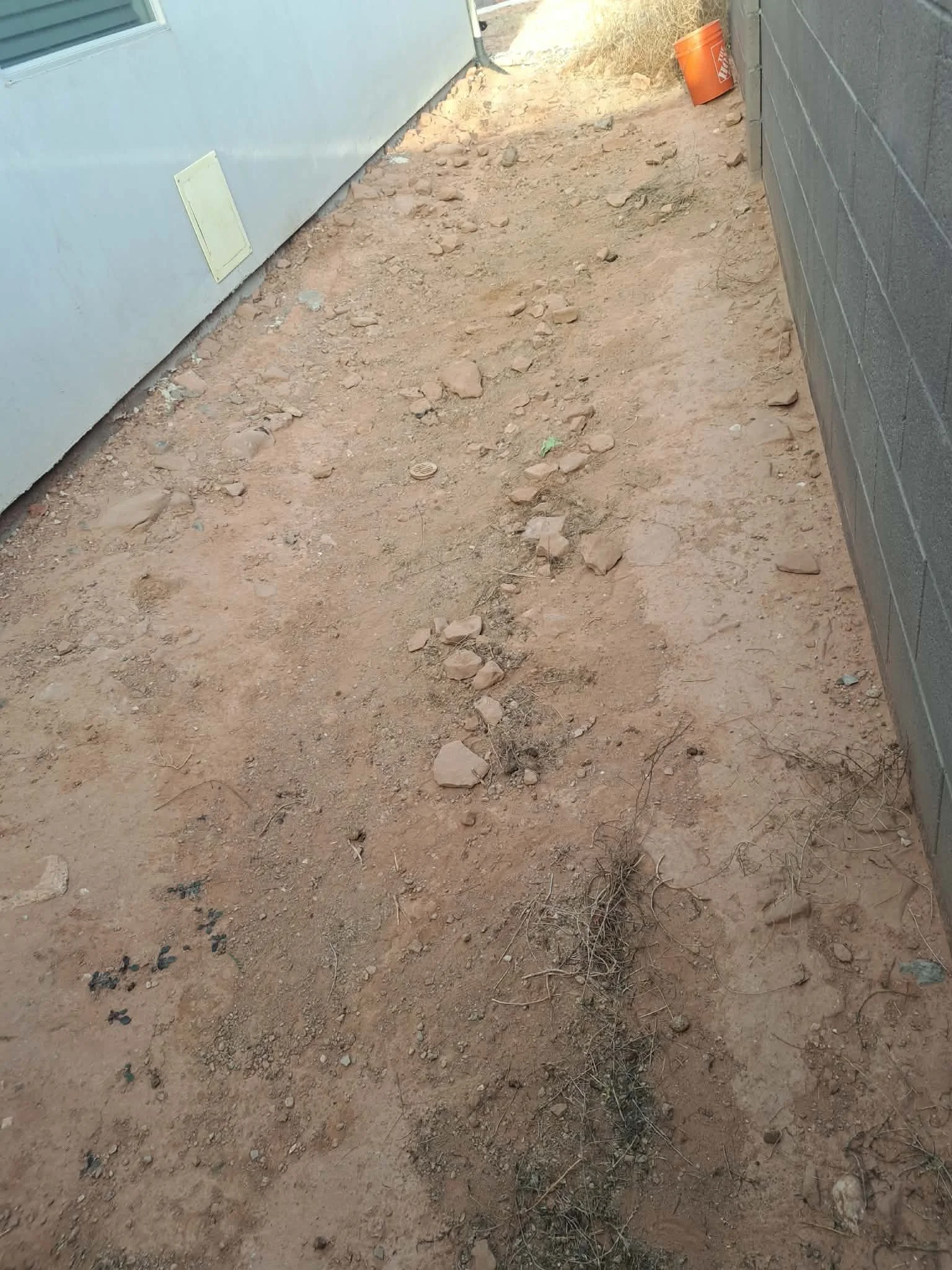 Outdoor construction site with dirt ground, small rocks, and sparse dry grass next to a building with a light gray wall and a darker gray wall.