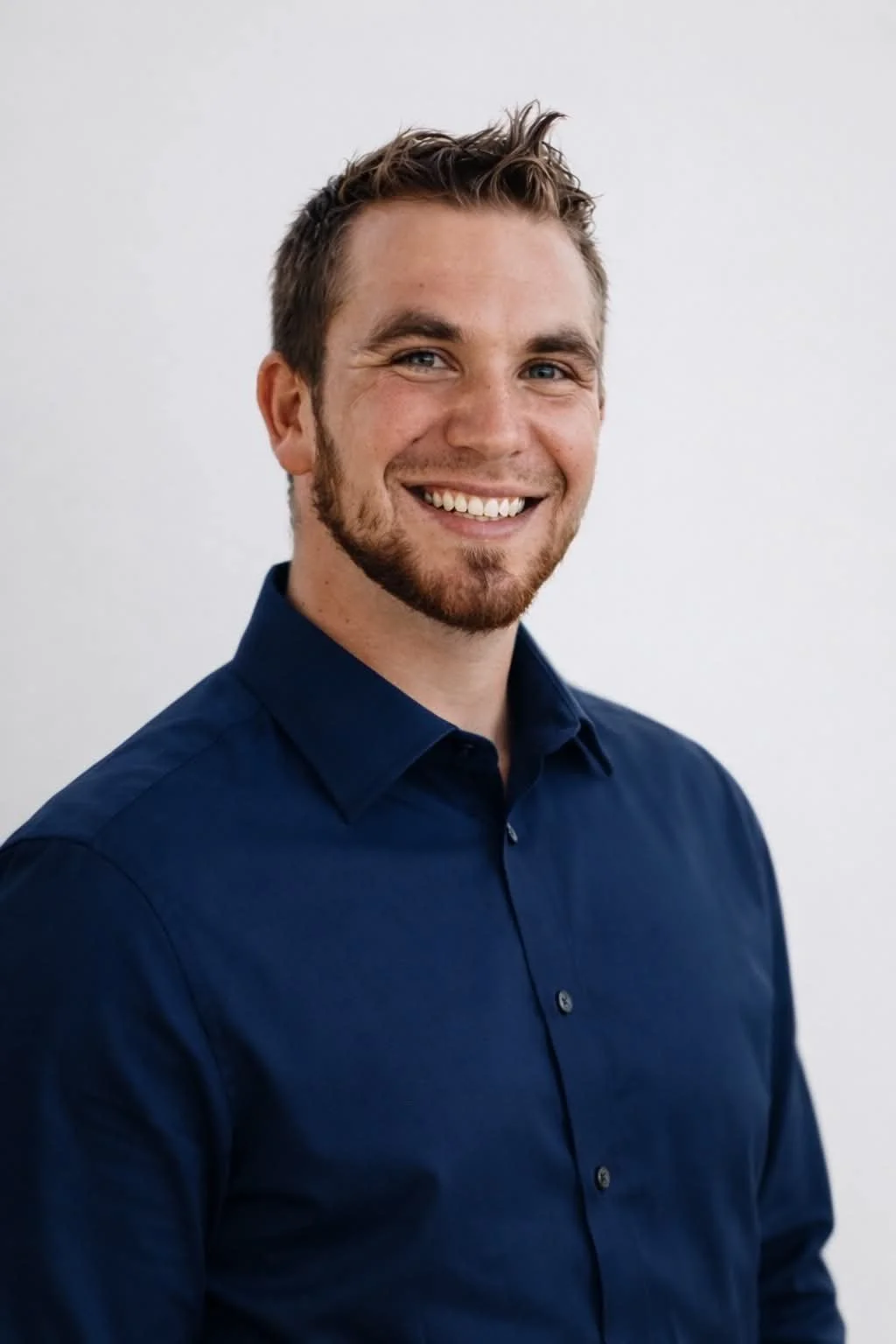 A smiling young man with short brown hair, blue eyes, and a beard, wearing a dark blue collared shirt, standing against a plain white background.