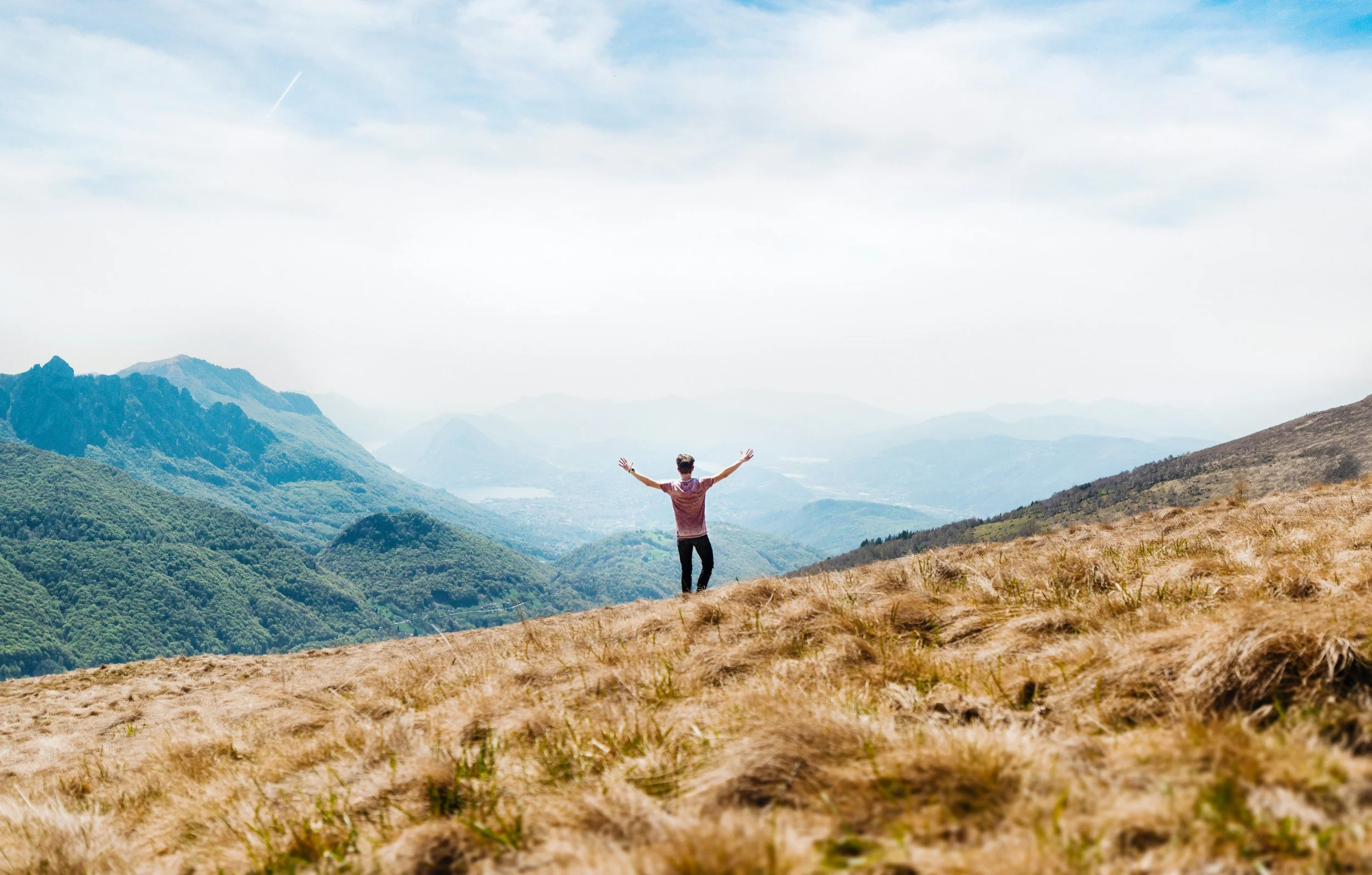 A person standing with arms outstretched on a grassy hillside overlooking mountains and a valley beneath a partly cloudy sky.