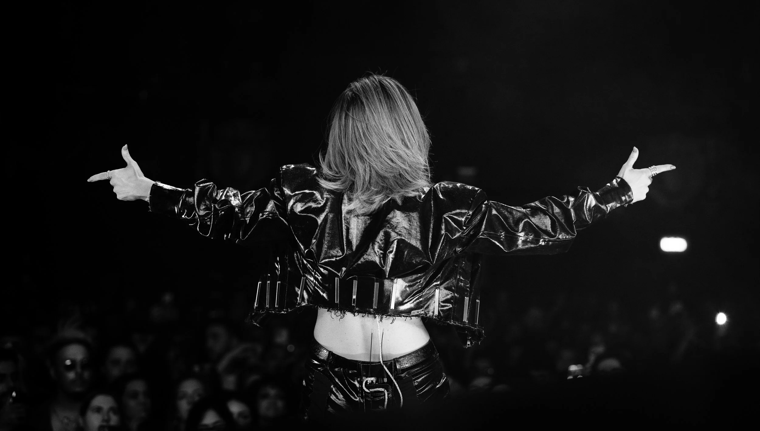 A woman with styled hair and a leather jacket standing with her back to the camera, arms outstretched, in front of a crowd during a performance.