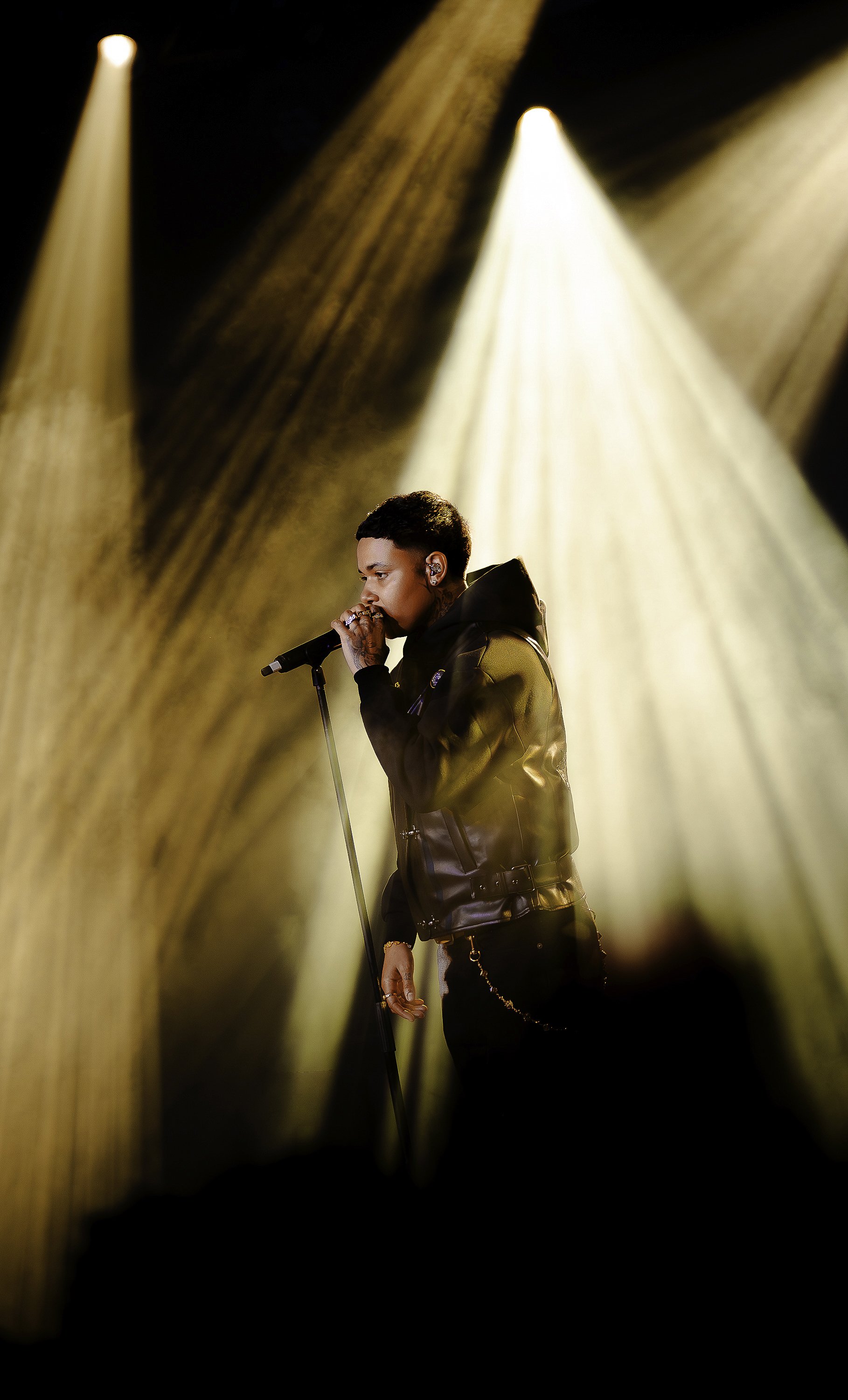 A male singer performs on stage, holding a microphone with both hands, under multiple bright spotlights.