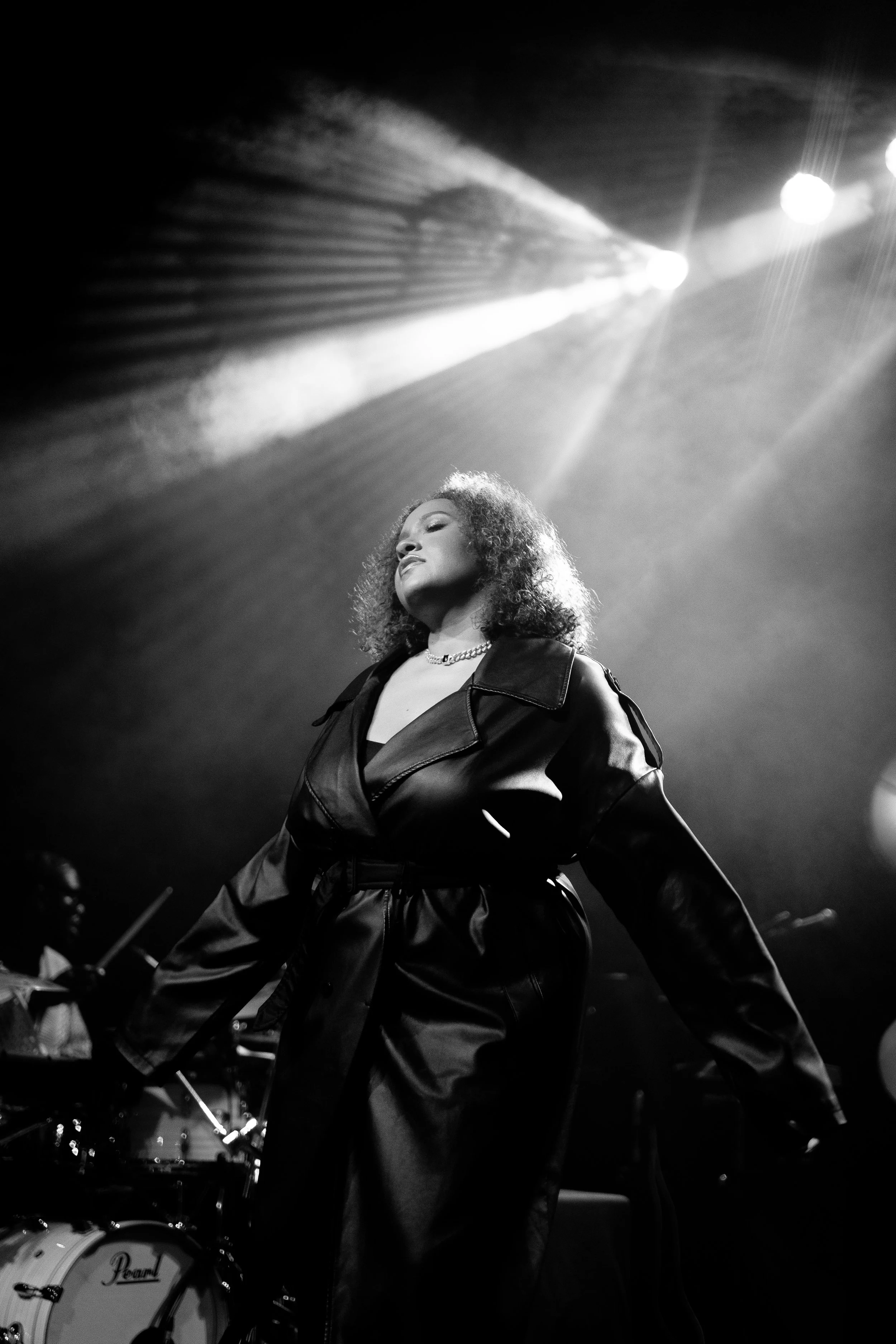 A woman with curly hair performs on stage under bright lights, wearing a leather jacket and a necklace, with a drum set in the background.