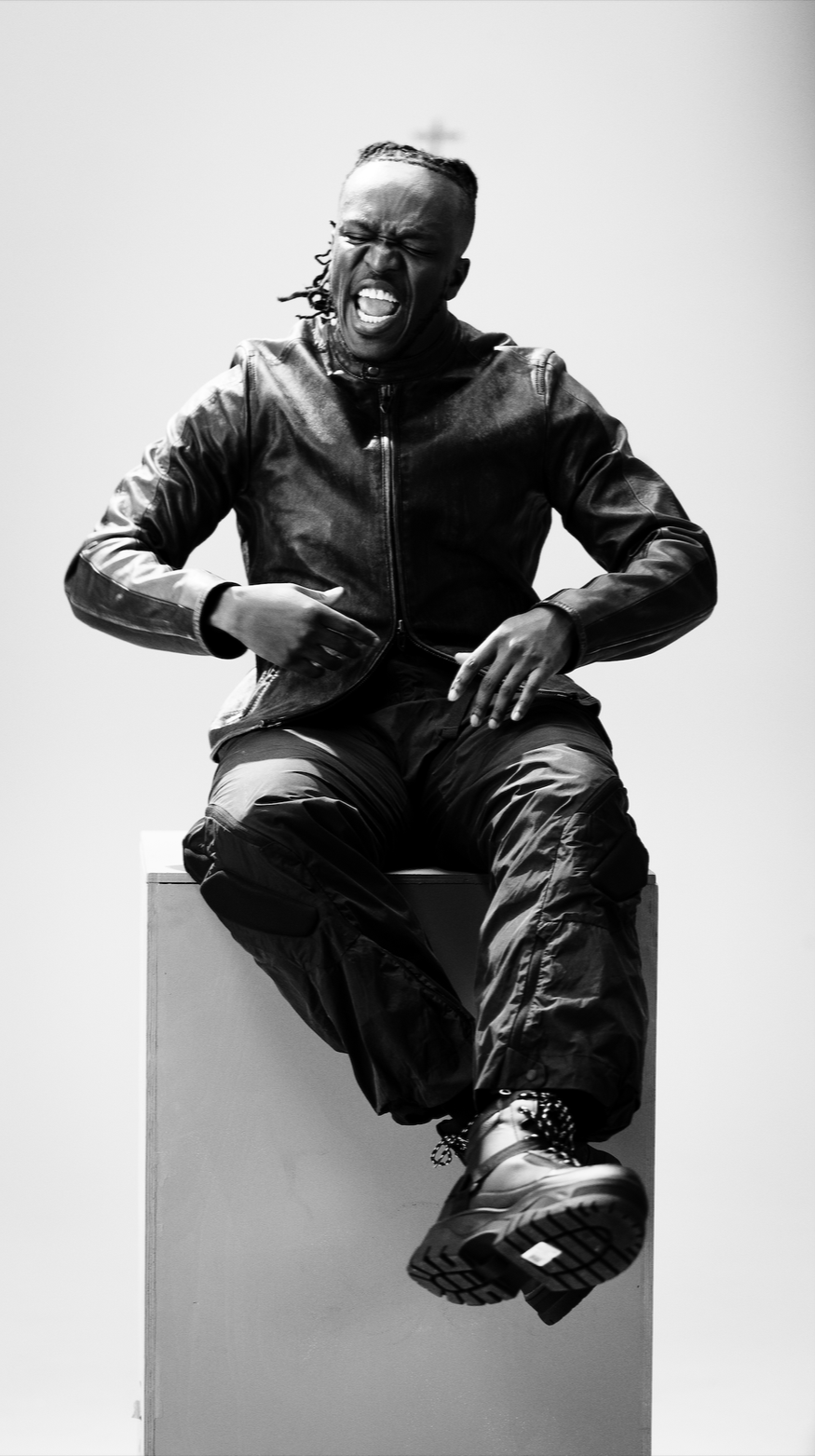 A person wearing a leather jacket, cargo pants, and sneakers, sitting on a box, with an open-mouthed expressive facial expression in front of a plain background.