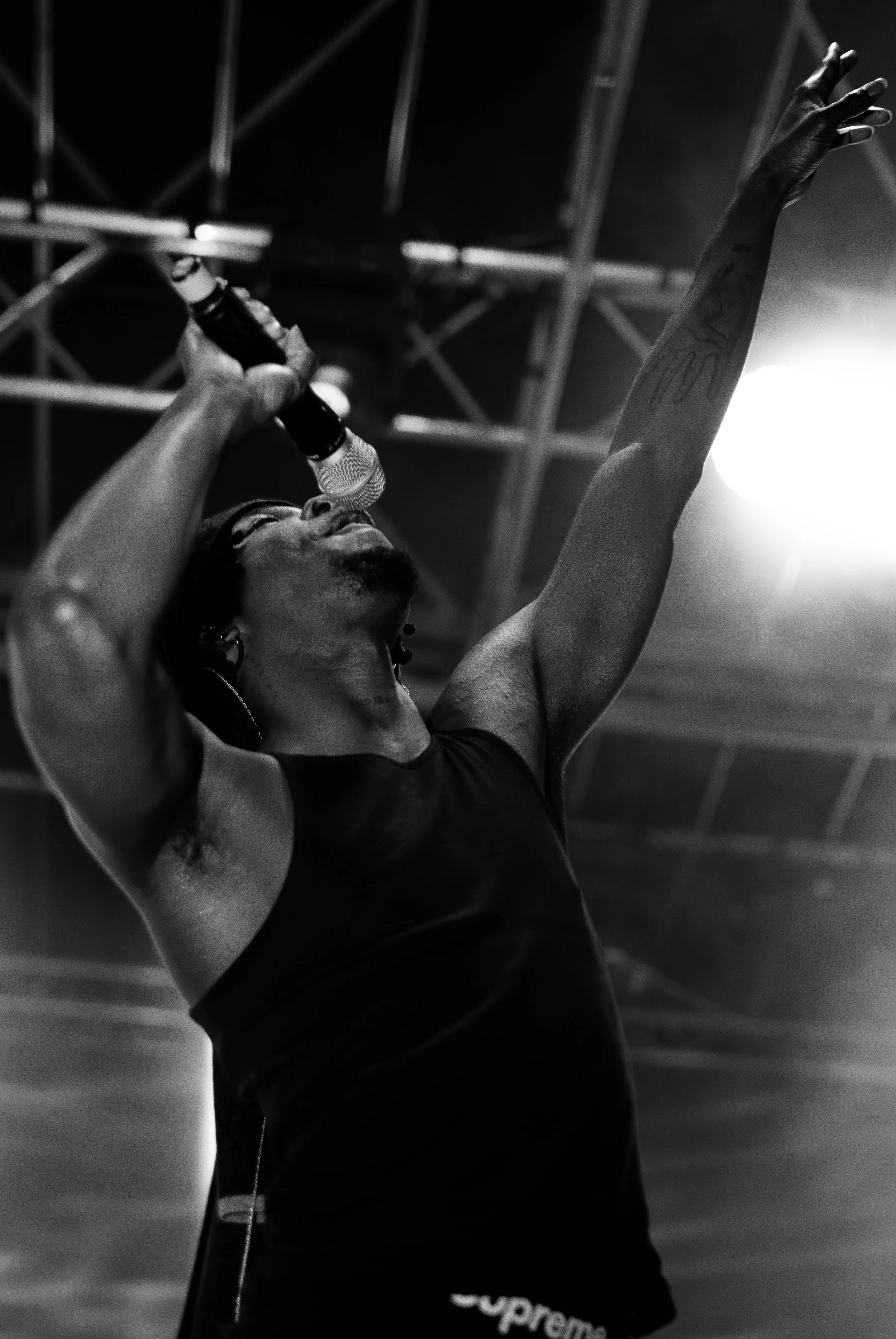A man with tattoos on his arm singing into a microphone with his right hand raised, in a dark indoor setting with visible ceiling beams, wearing a sleeveless shirt and black shorts with a 'Supreme' logo.