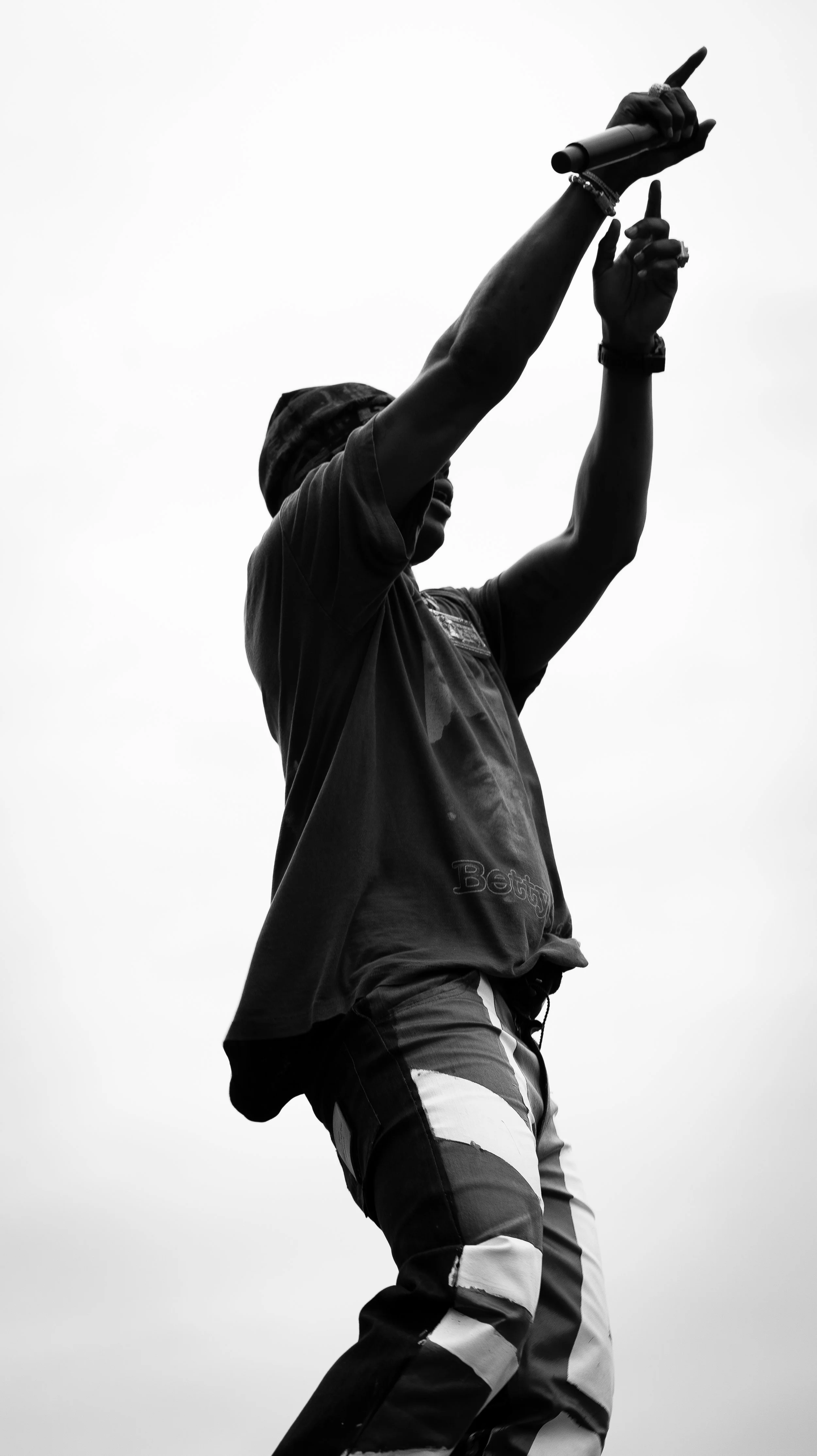 Black and white photo of a person singing into a microphone with one hand raised, wearing a beanie and patterned pants.